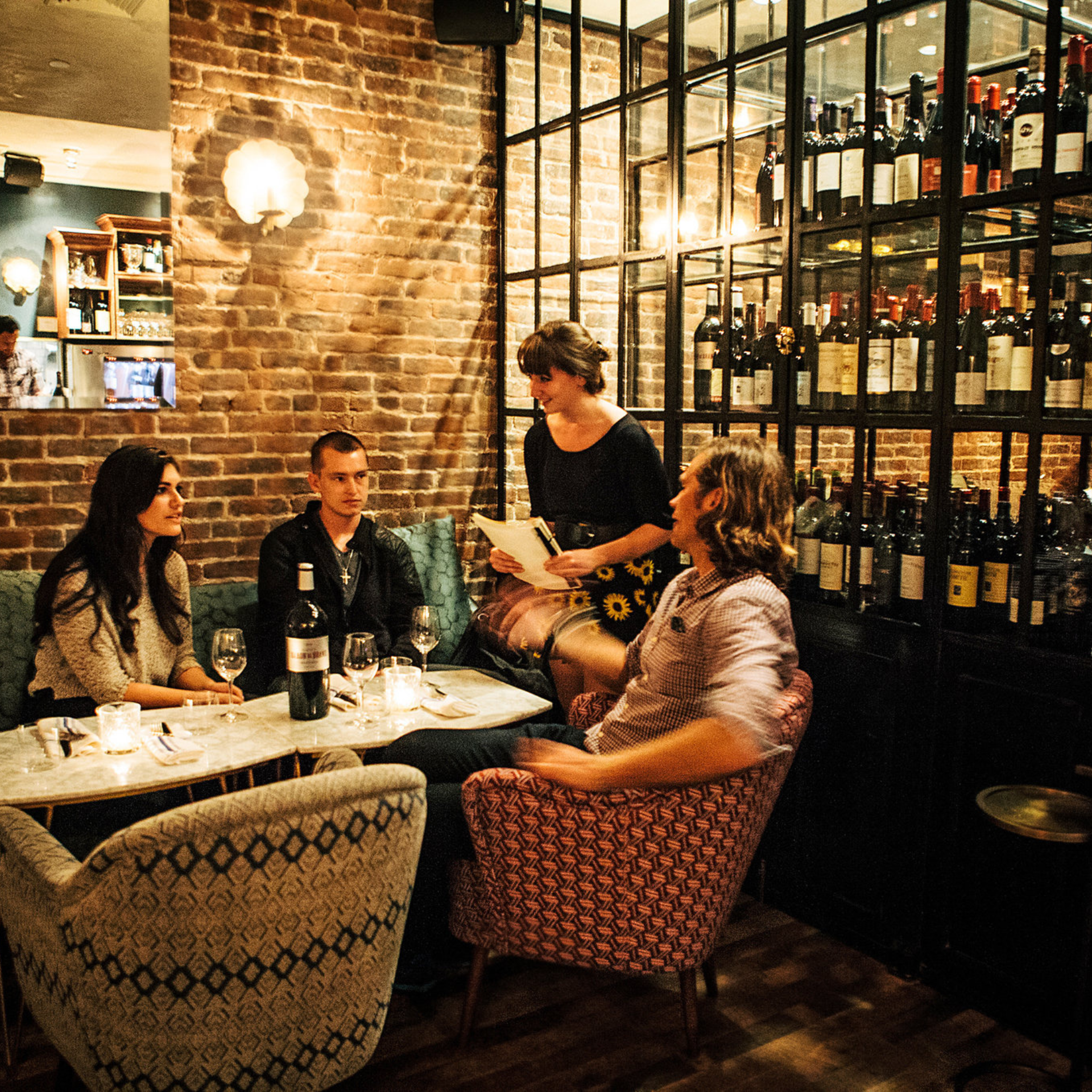 Upscale wine bar with brick walls, soft lighting, wine cabinet, and guests chatting at a candlelit table with a server nearby