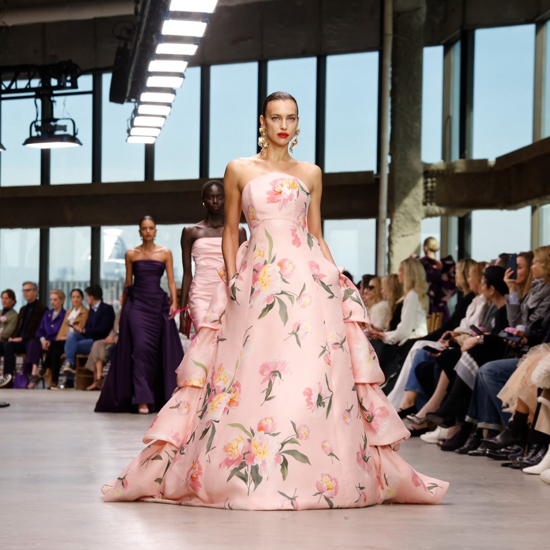 Model in floral gown walking fashion runway with audience and natural light