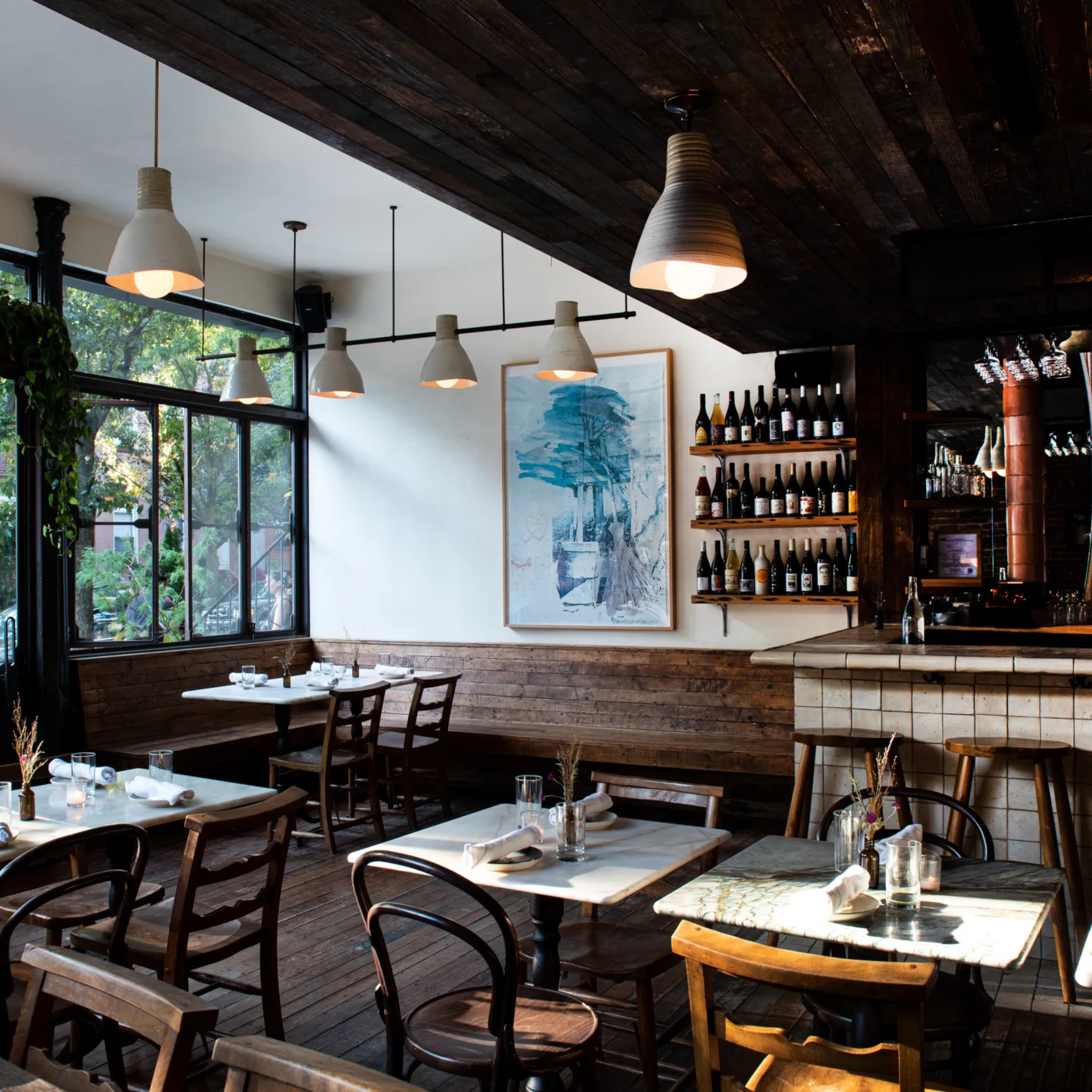 Rustic restaurant with wood floors, marble tables, pendant lights, wine shelf, and warm natural light from large windows