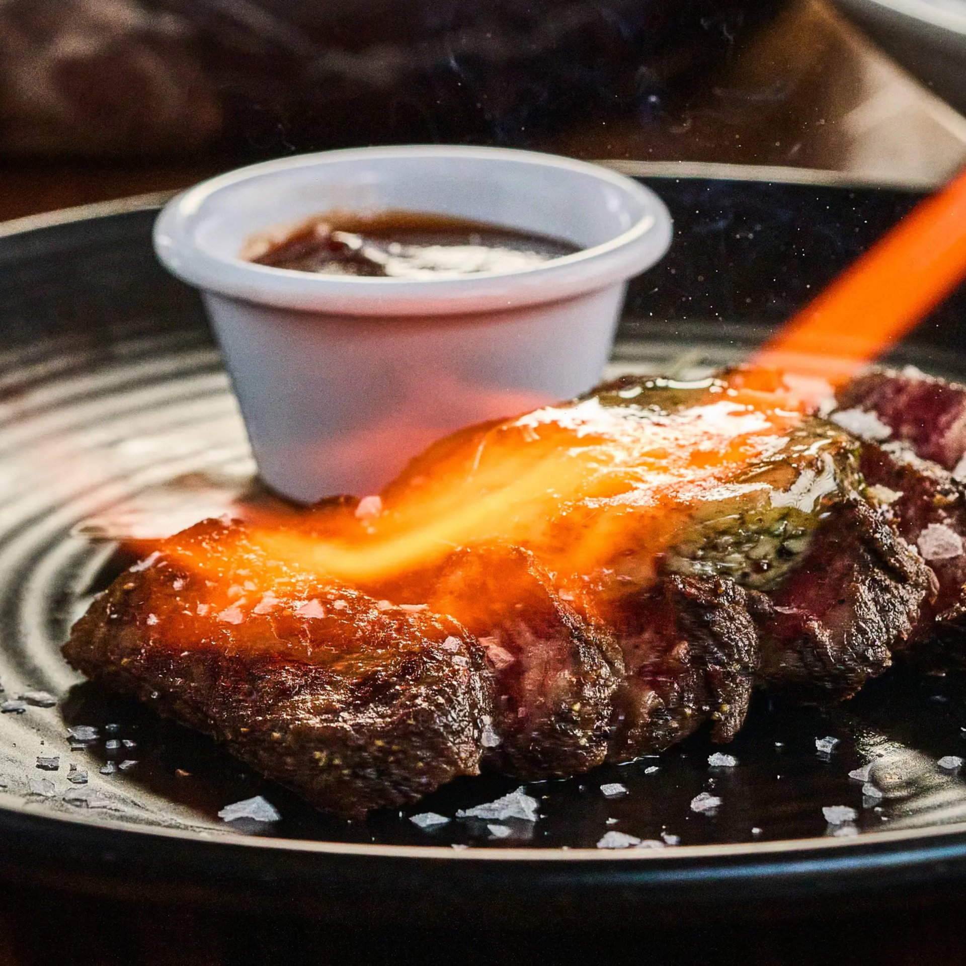 Blowtorch searing steak with salt and sauce ramekin