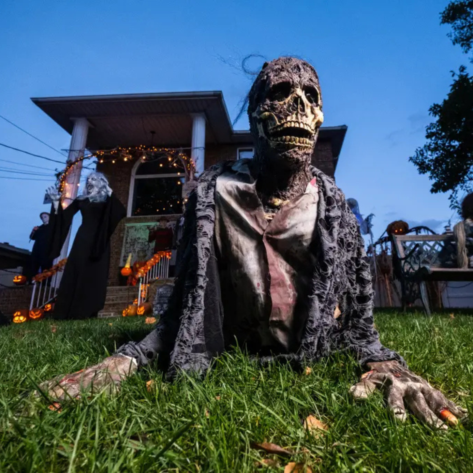 Creepy zombie decoration crawling on grass with skeleton face, in front of a house decorated with Halloween lights and props