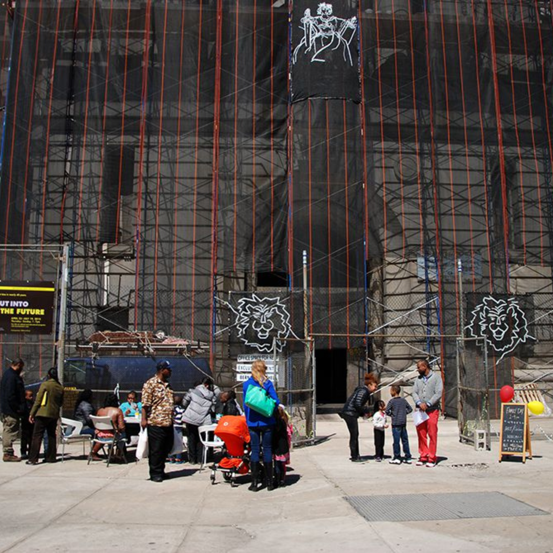 People gather outside a building under construction with lion art, banners, tables, and balloons in front