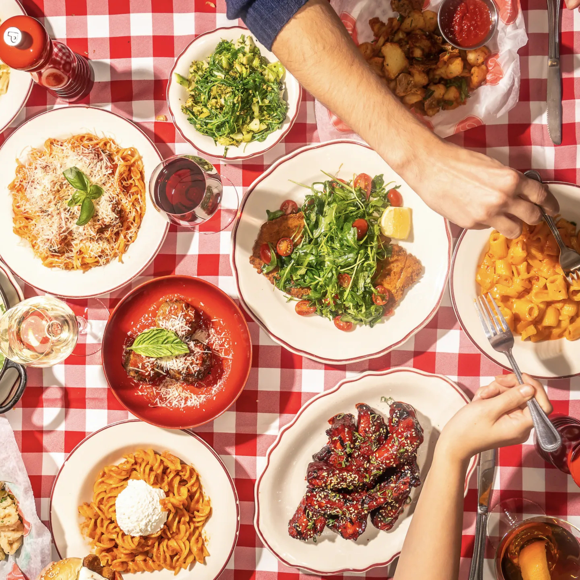 Red and white checkered table with Italian dishes: spaghetti, meatballs, rigatoni, salad, chicken wings, and wine glasses