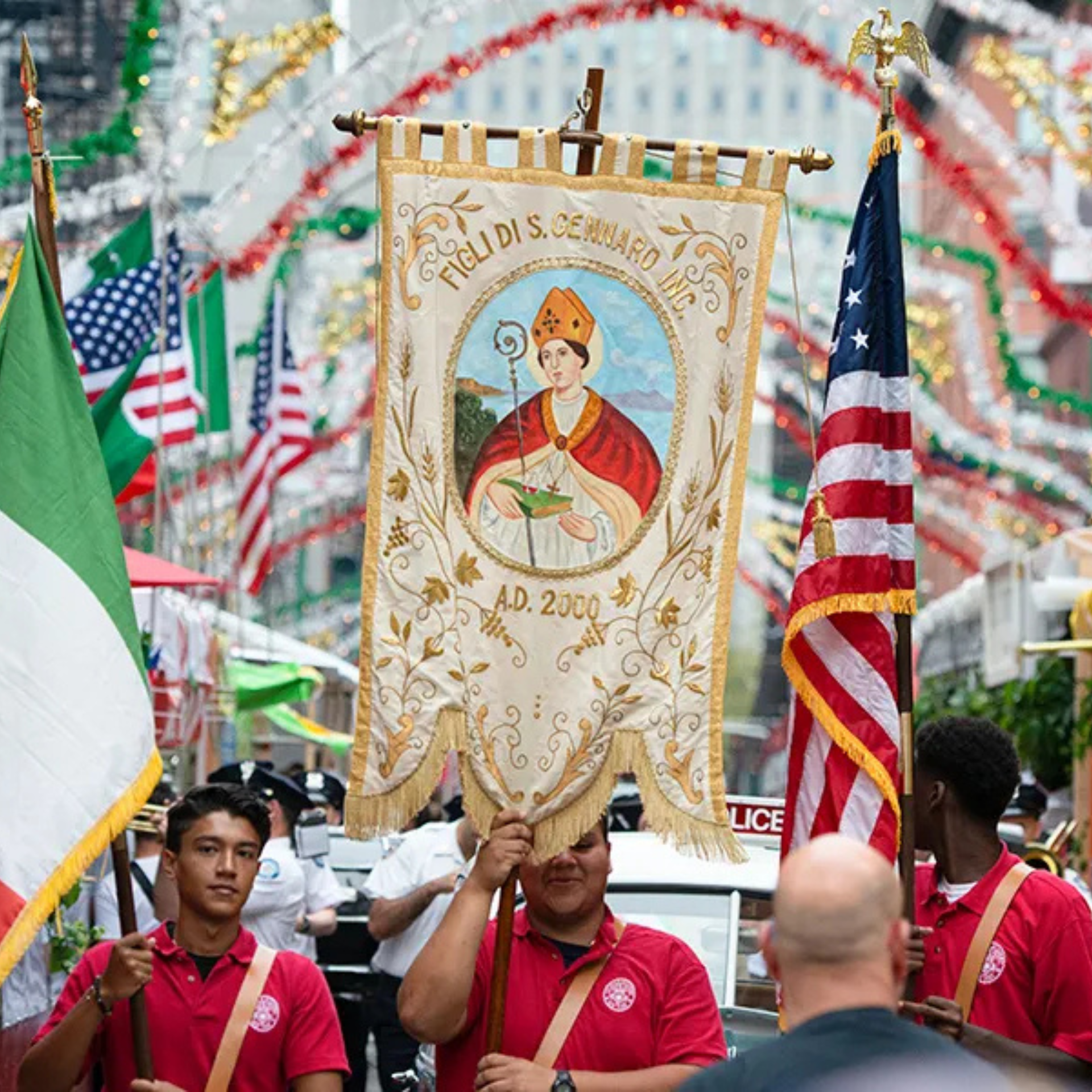 Parade participants in red shirts hold ornate banner; American and Italian flags wave amid festive city crowd