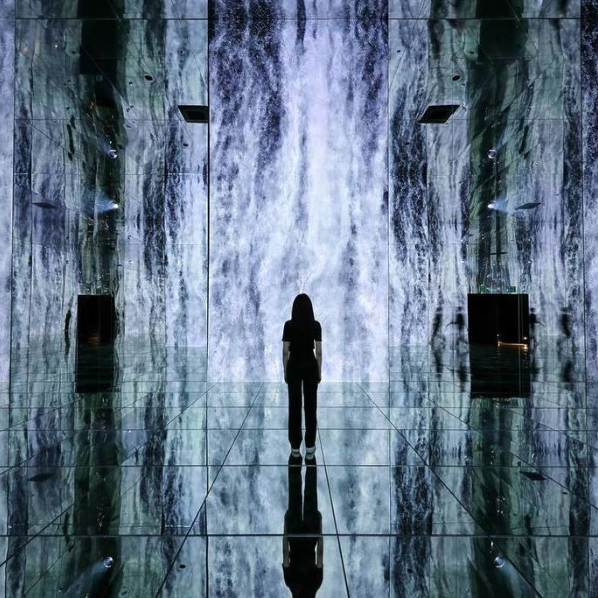 Person stands in a mirrored digital art installation, silhouetted against a cascading waterfall projection that reflects across the glossy floor and walls