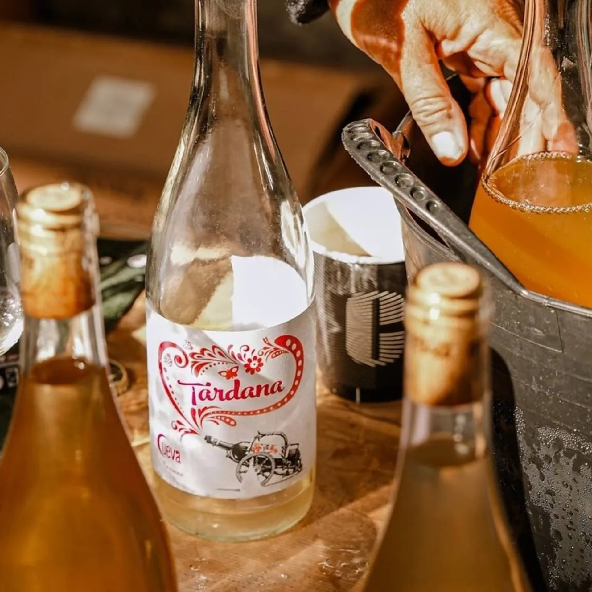 Bottles of light-colored wine or cider displayed on a wooden surface, with one labeled “Tardana” being placed into a black ice bucket during a tasting or serving setup