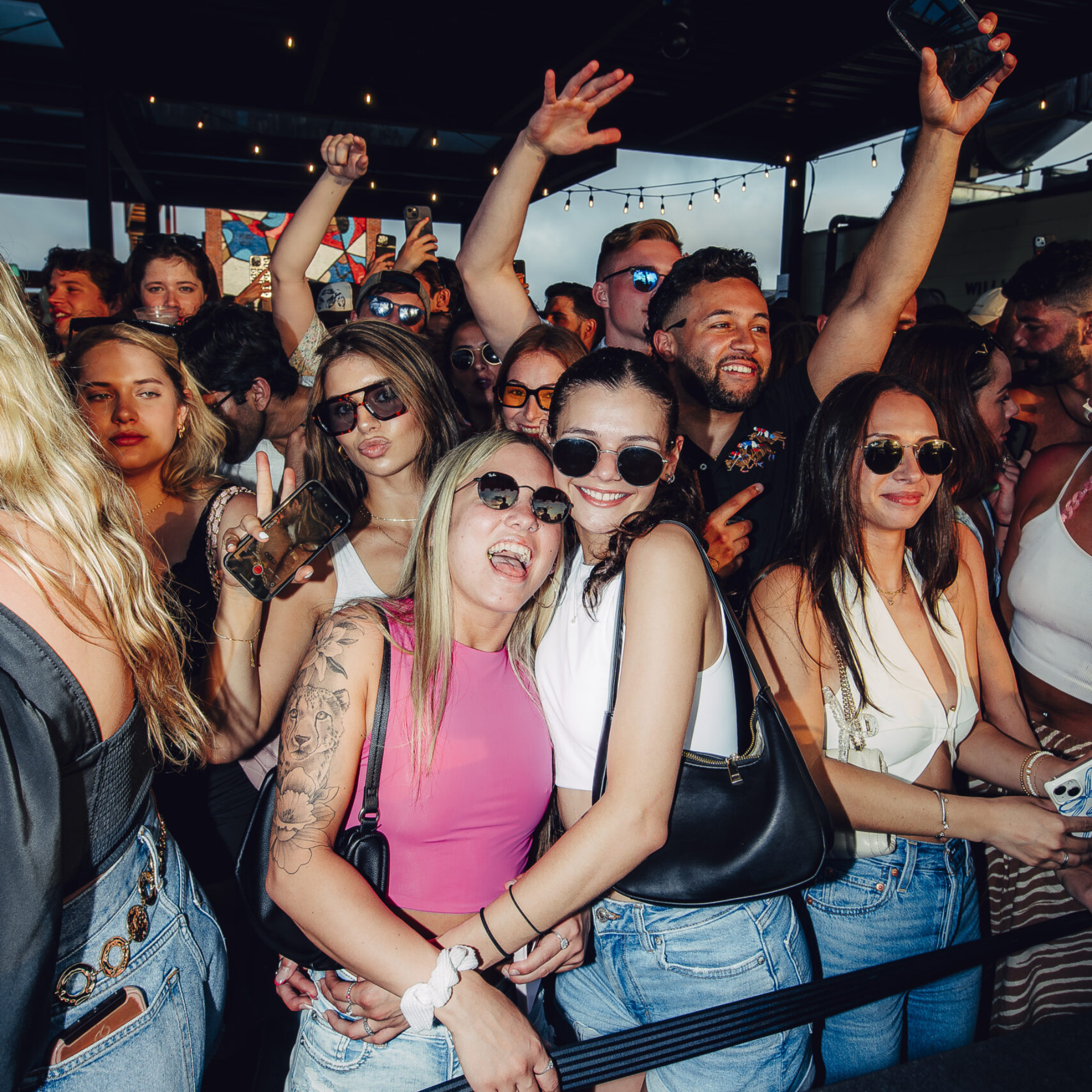 Crowd of young adults enjoying an outdoor summer party, smiling and posing in casual outfits under string lights