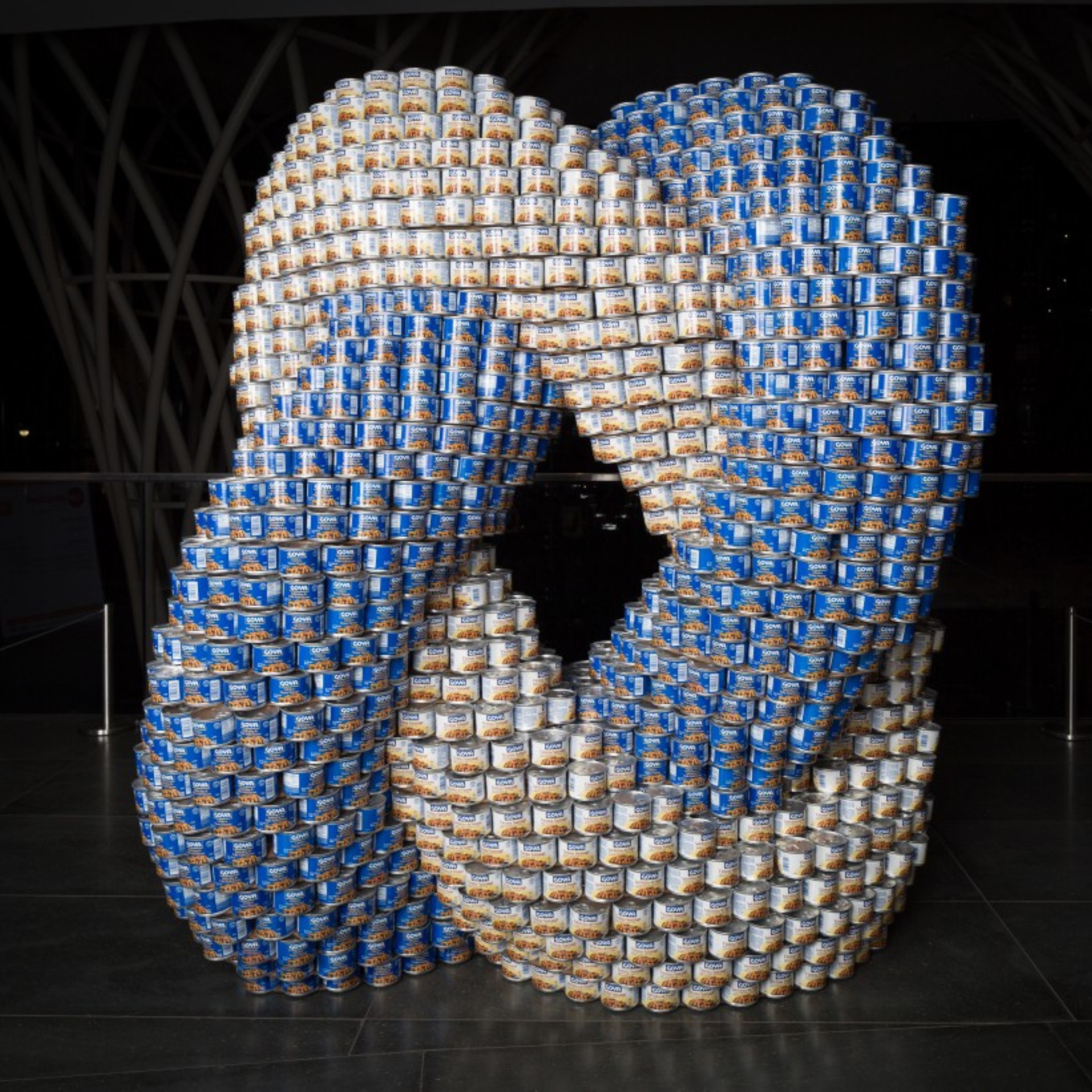 Intricate sculpture made entirely of stacked canned goods forms a twisted torus shape, using blue and white labels to create a striking visual centerpiece—likely part of a charitable art installation