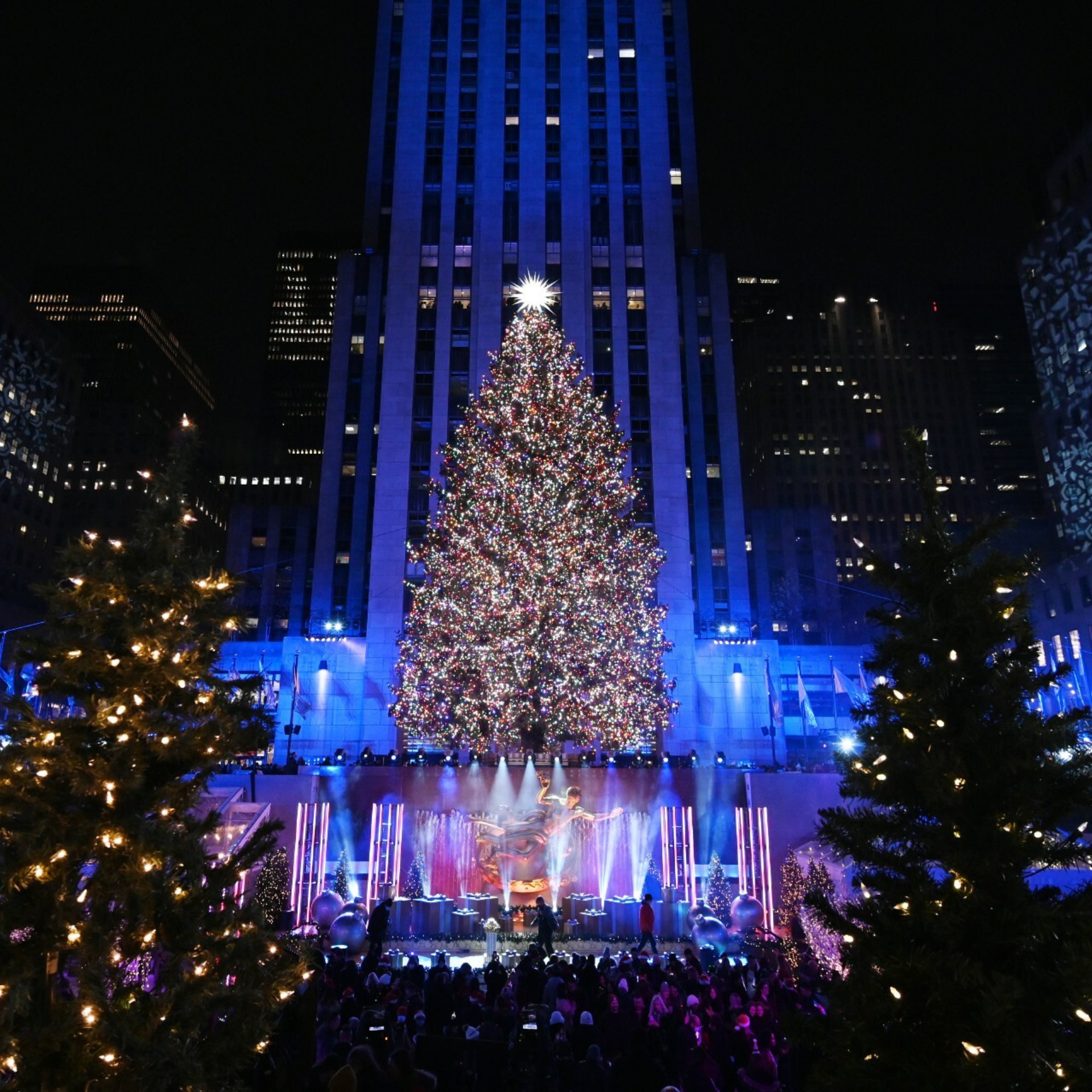 Rockefeller Center’s Christmas Tree glows with multicolored lights and a star topper, surrounded by festive crowds and city towers