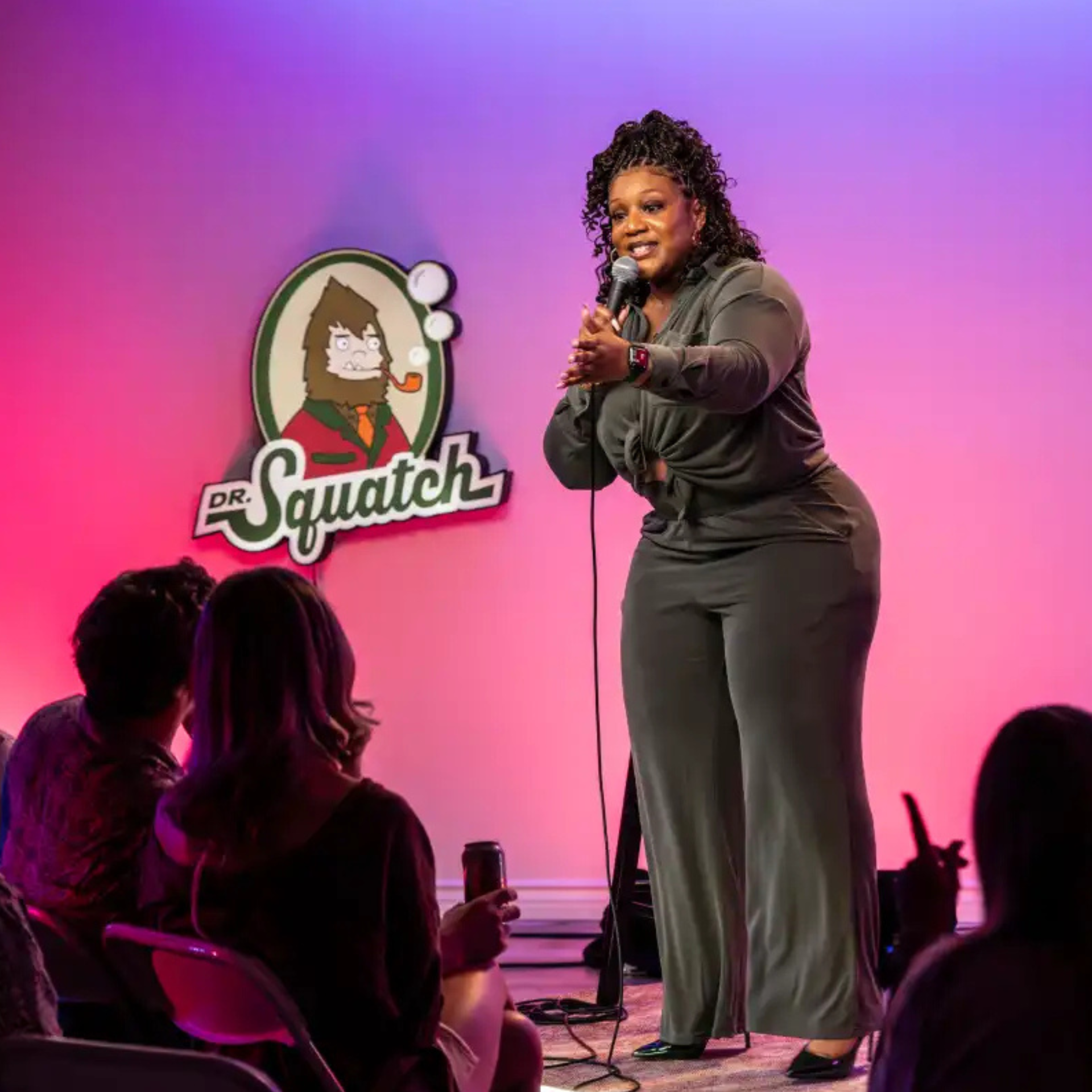 Stand-up comic performs under pink-purple lights as audience laughs; Dr. Squatch logo glows behind in lively event setting