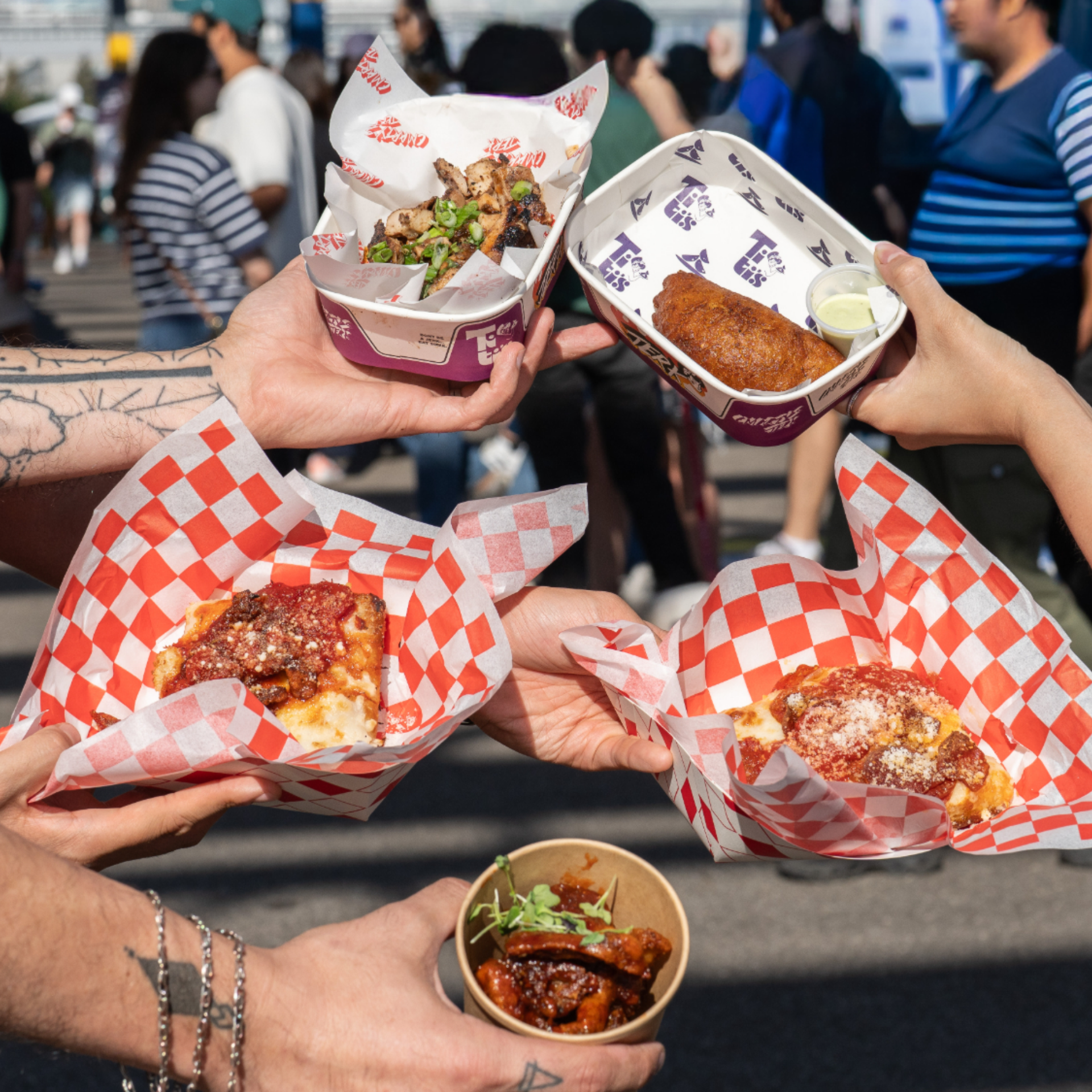 Hands holding pizza, wings, and fried foods at outdoor market