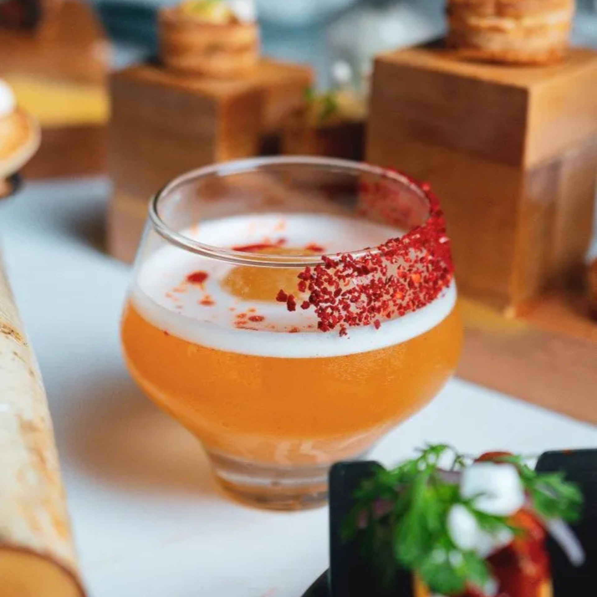 Cocktail in a rounded glass with a frothy top and red seasoning on the rim, served alongside gourmet dishes in an upscale dining setting