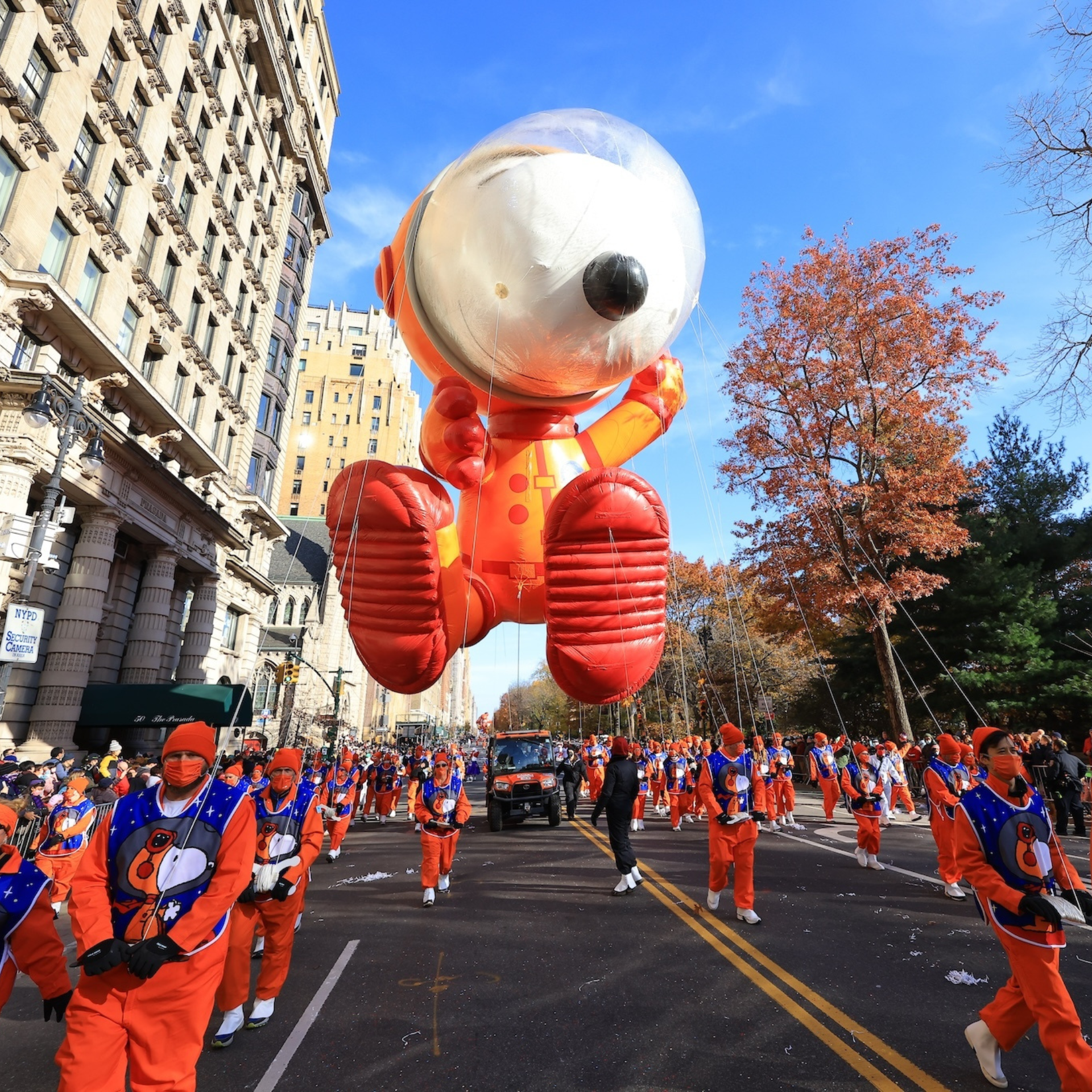 Astronaut Snoopy balloon floats above a NYC street during a festive parade, guided by participants in matching orange suits beneath a clear autumn sky