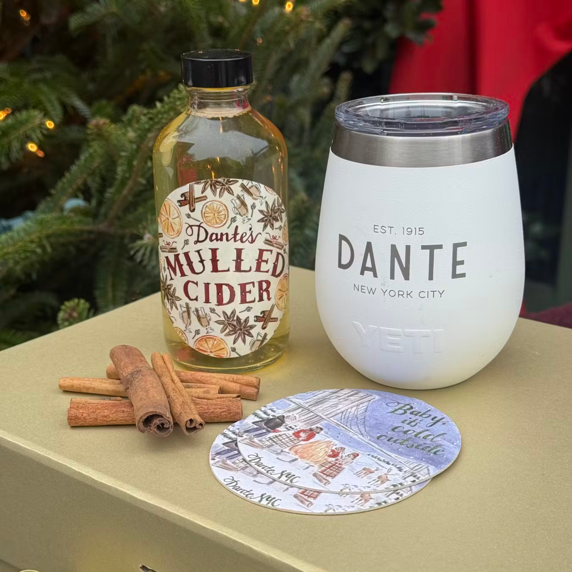 Festive setup featuring Dante’s Mulled Cider, cinnamon sticks, and a branded tumbler beside a “Baby it’s cold outside” coaster