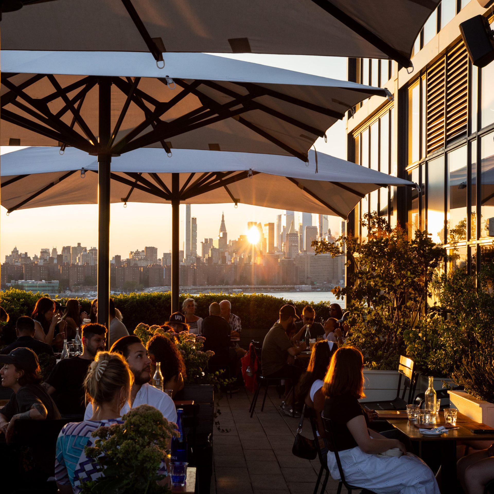 Rooftop bar buzzes at sunset with diners under umbrellas, NYC skyline glowing behind glass buildings and golden-lit skyscrapers