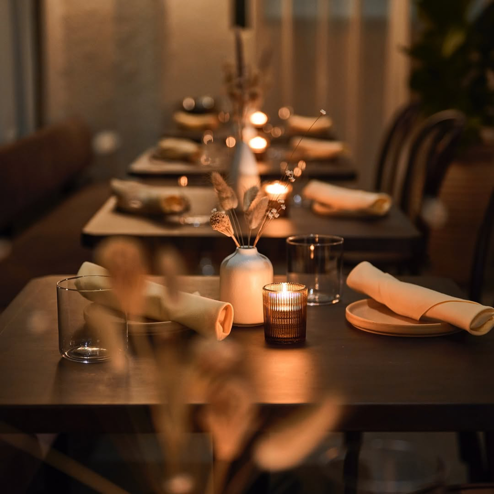 Warmly lit dining setup with rolled beige napkins, wooden plates, dried flowers, and candles on a long table