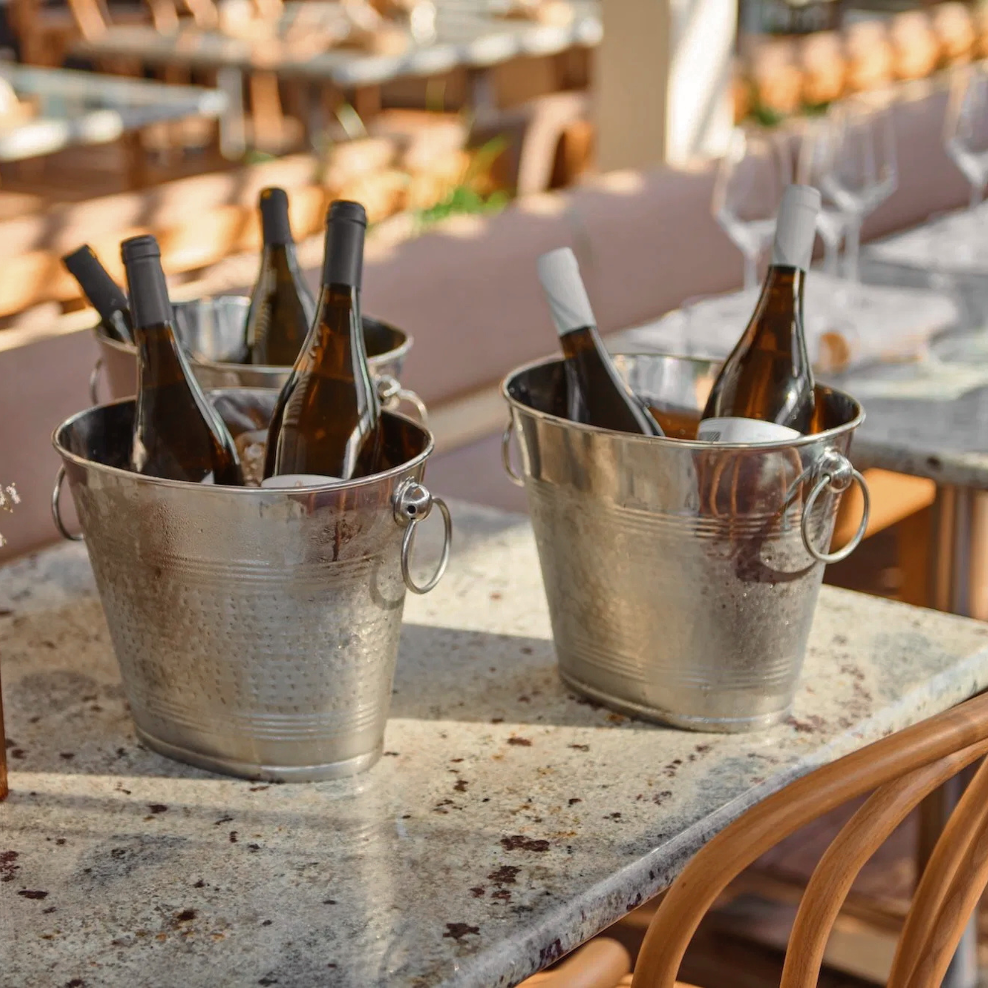 Two metal buckets filled with wine bottles on an outdoor table, with chairs and glasses set for dining in the background
