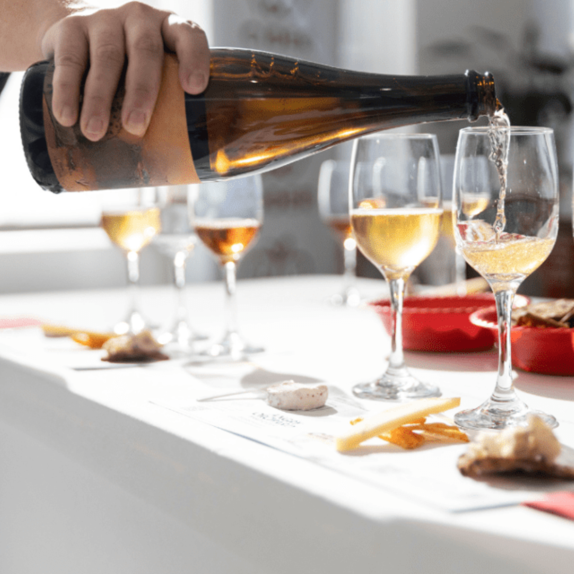 Hand pours golden wine into glasses during a tasting, with small bites like cheese and dried fruit arranged on a white surface in bright daylight