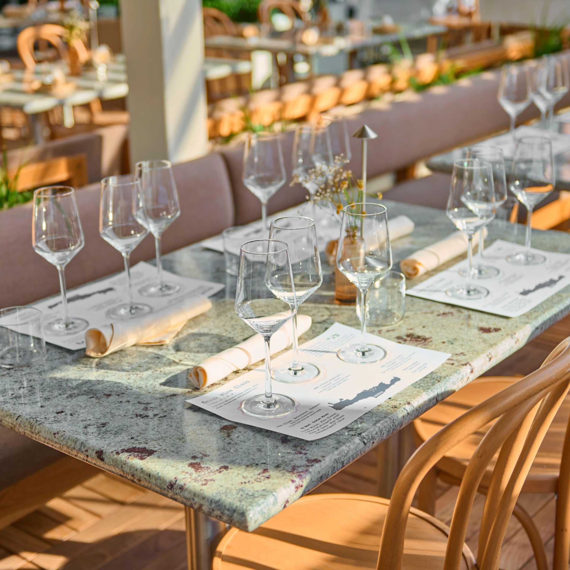 Elegant table set for a wine tasting, featuring multiple glasses, rolled napkins, and printed menus on a speckled stone surface in a sunlit venue