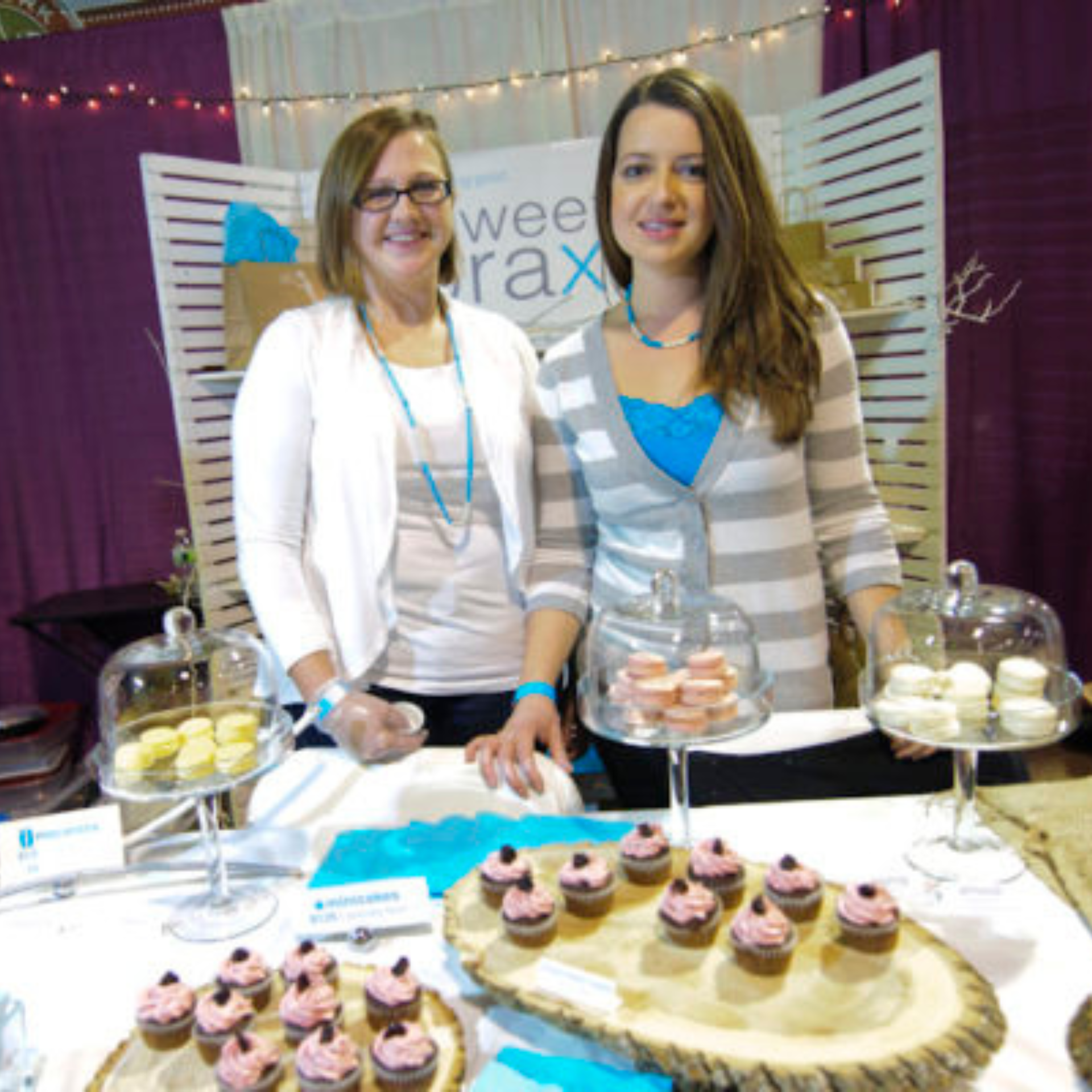 Two women stand behind a bakery booth displaying pink-frosted cupcakes and colorful macarons under glass domes, promoting their treats at a festive market