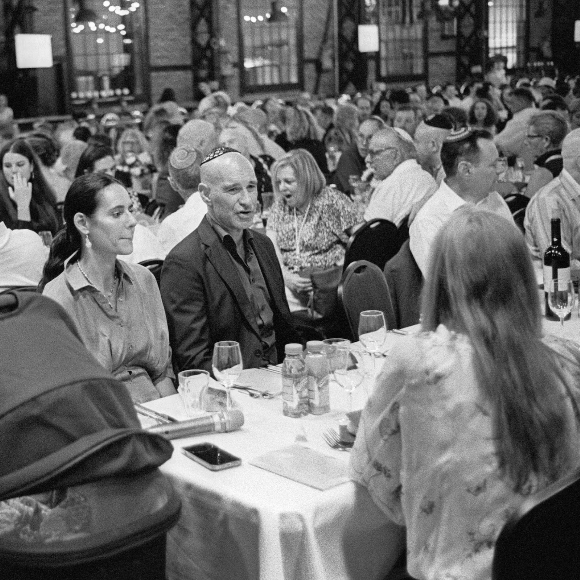 Guests gather for a communal meal in a festive indoor setting, with many wearing kippahs and seated at tables set with wine, drinks, and conversation