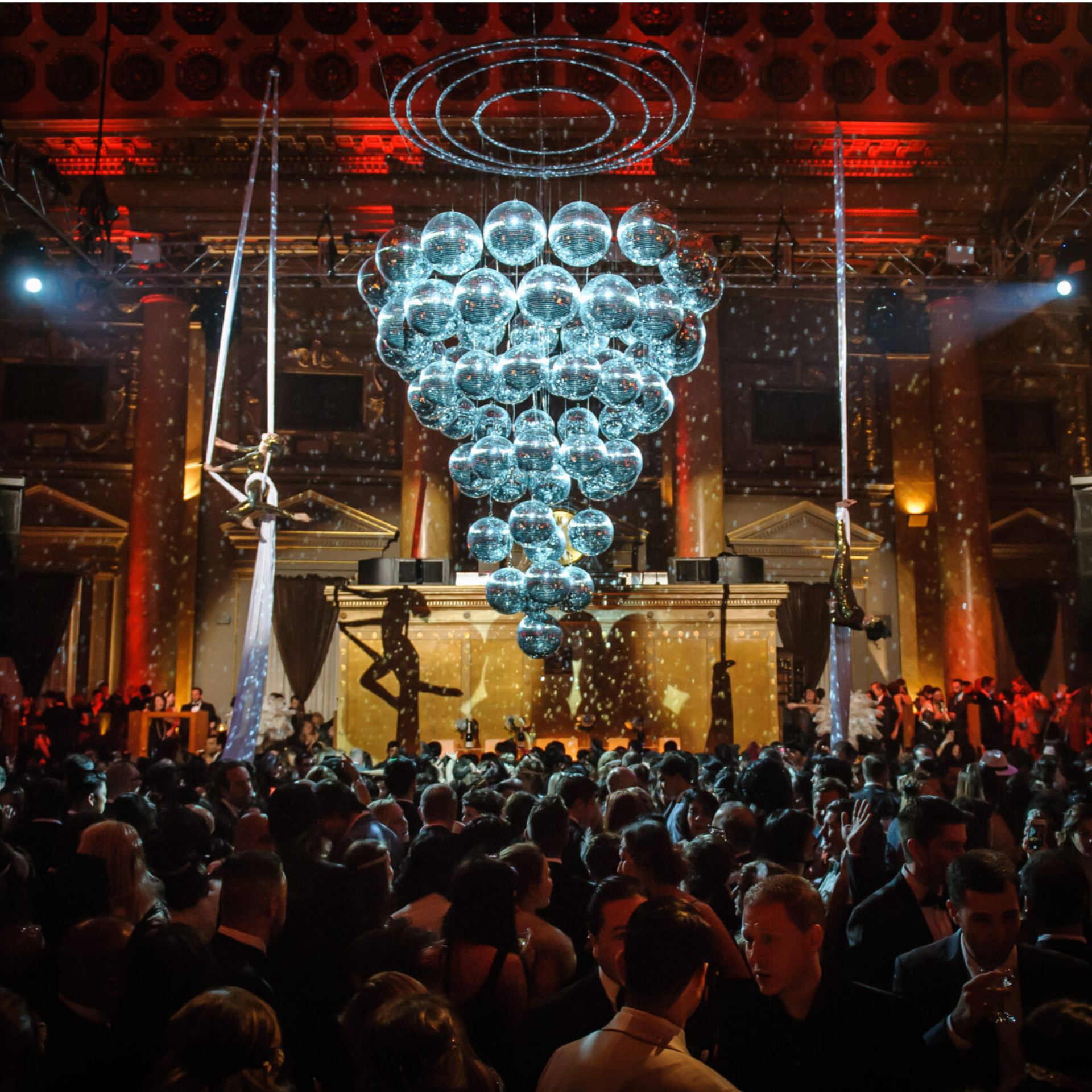 Formal crowd in grand ballroom with aerial performers and chandelier spheres