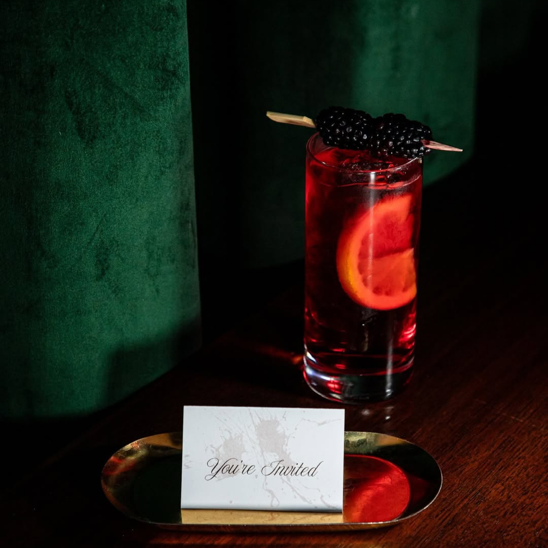 Red cocktail with blackberries and orange slice beside a “You’re Invited” card on a luxe velvet backdrop