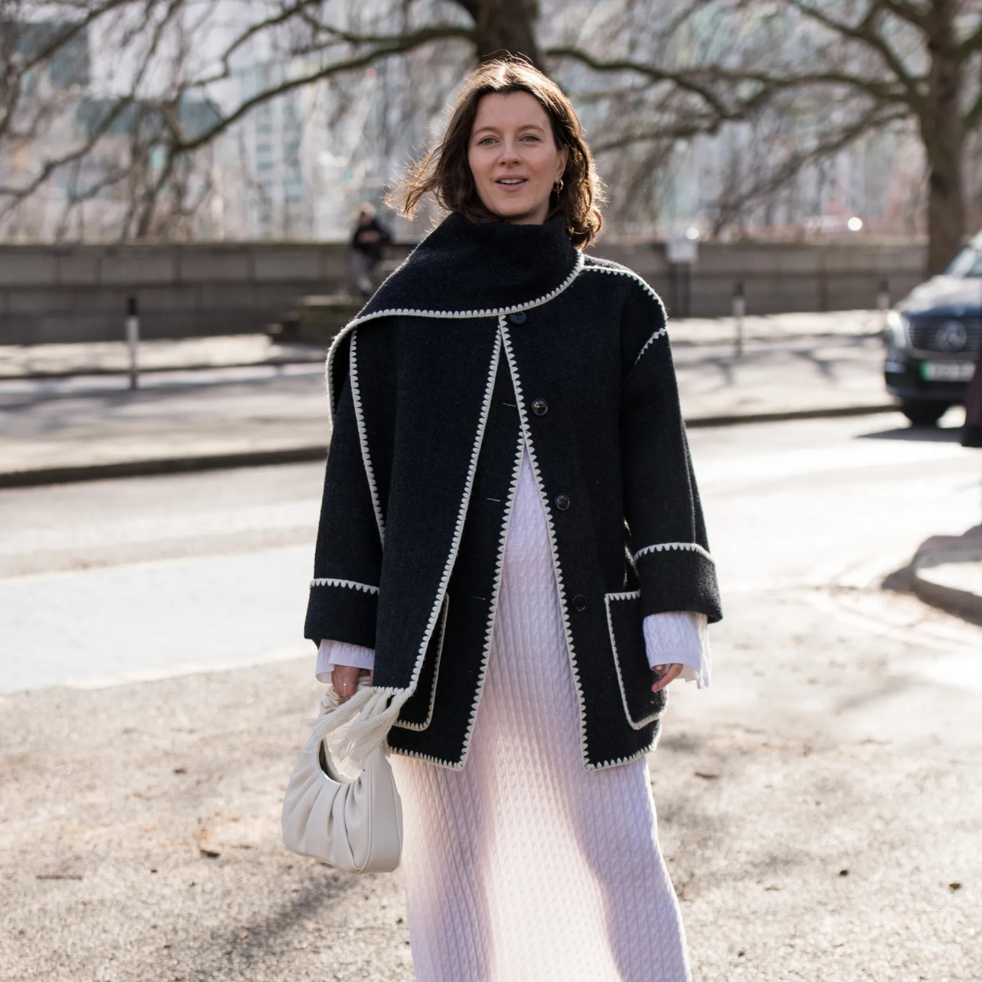 Stylish city look: black coat with white trim, textured white skirt, and handbag. Leafless trees hint at late fall fashion