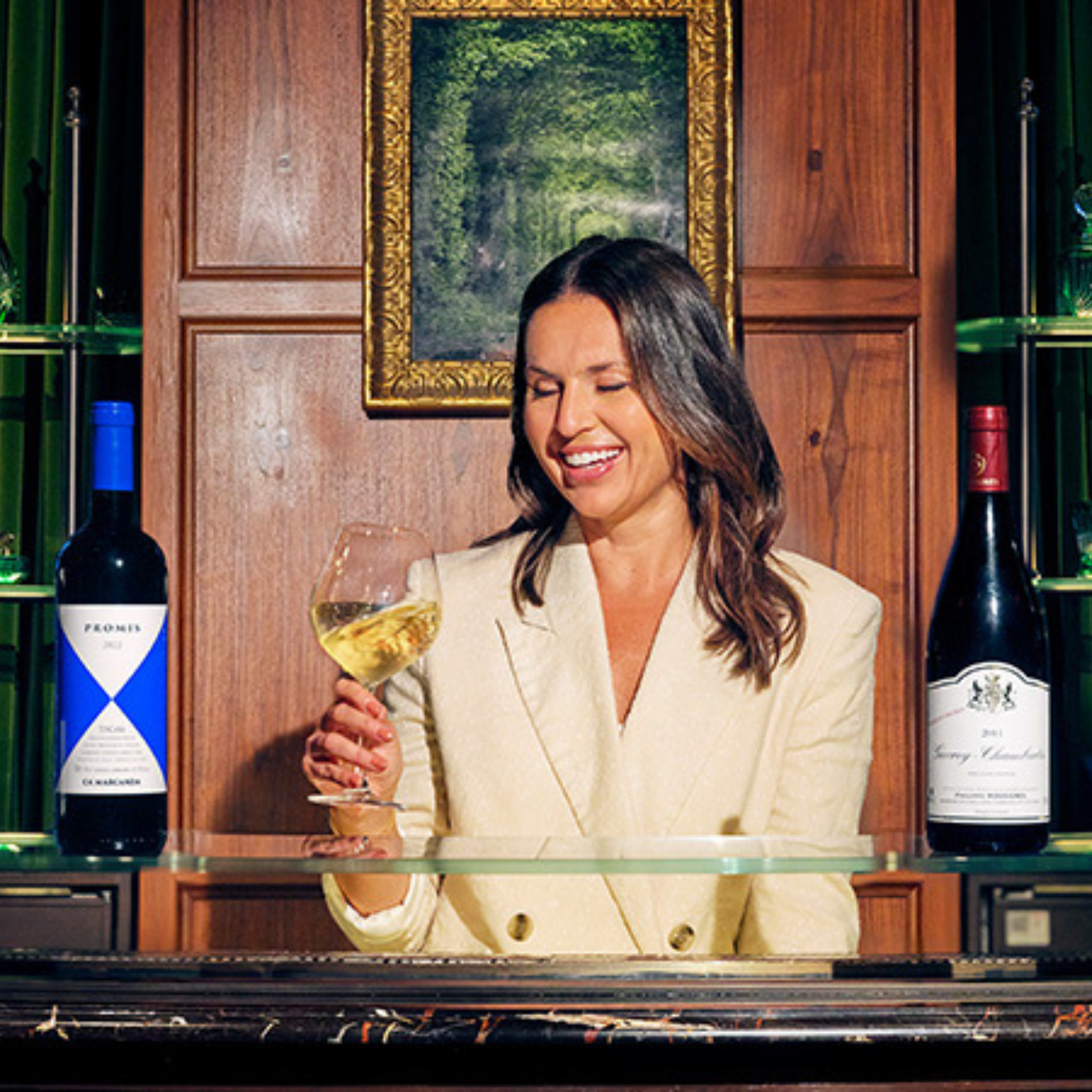 Woman in a cream blazer holding a glass of white wine, standing behind a bar with two wine bottles and a wooden cabinet in the background