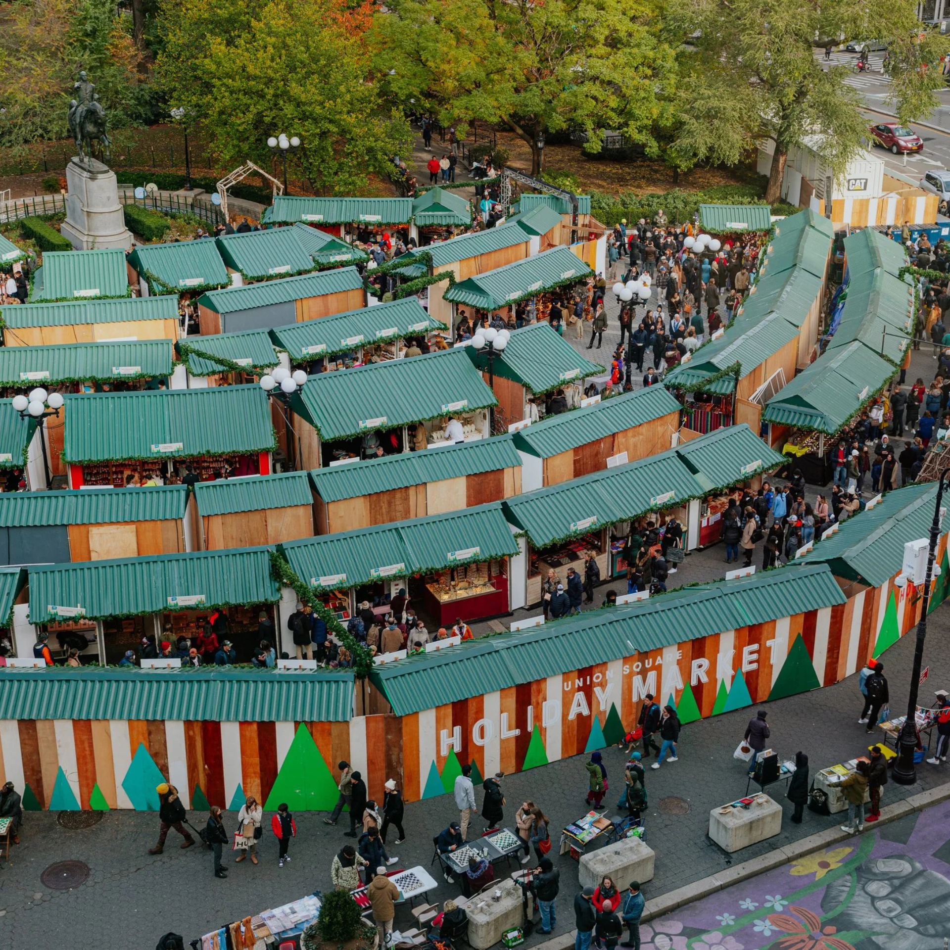 Aerial view of the Union Square Holiday Market in NYC, with green-roofed stalls winding through a festive plaza filled with shoppers and autumn trees