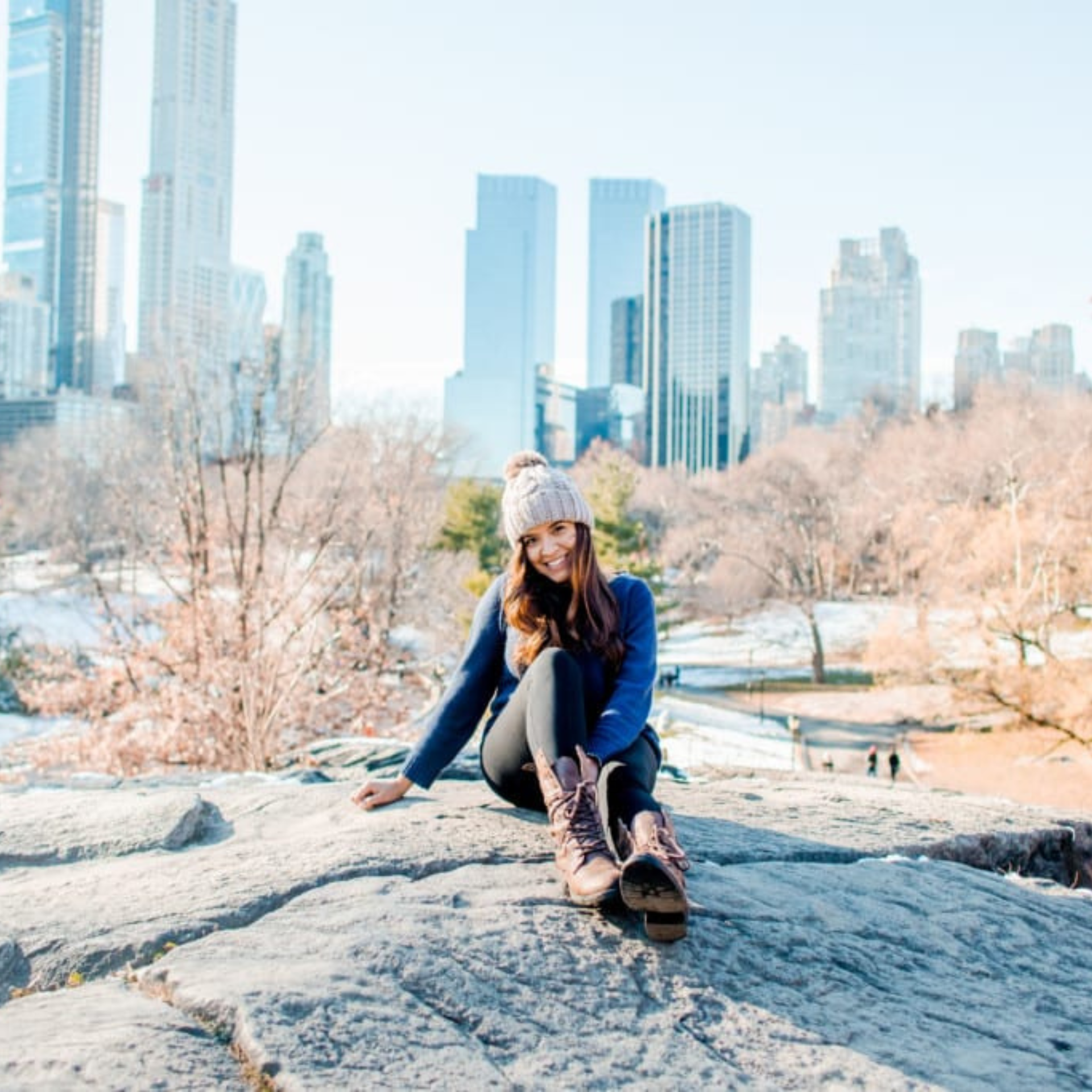 Person in knit hat and boots sits on Central Park rocks, smiling with NYC skyline behind bare trees on a crisp winter day