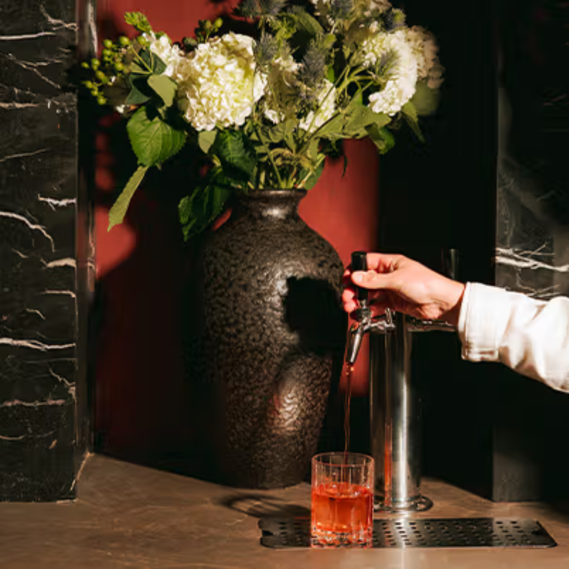 Hand pouring red drink from tap beside black vase with white flowers on marble counter