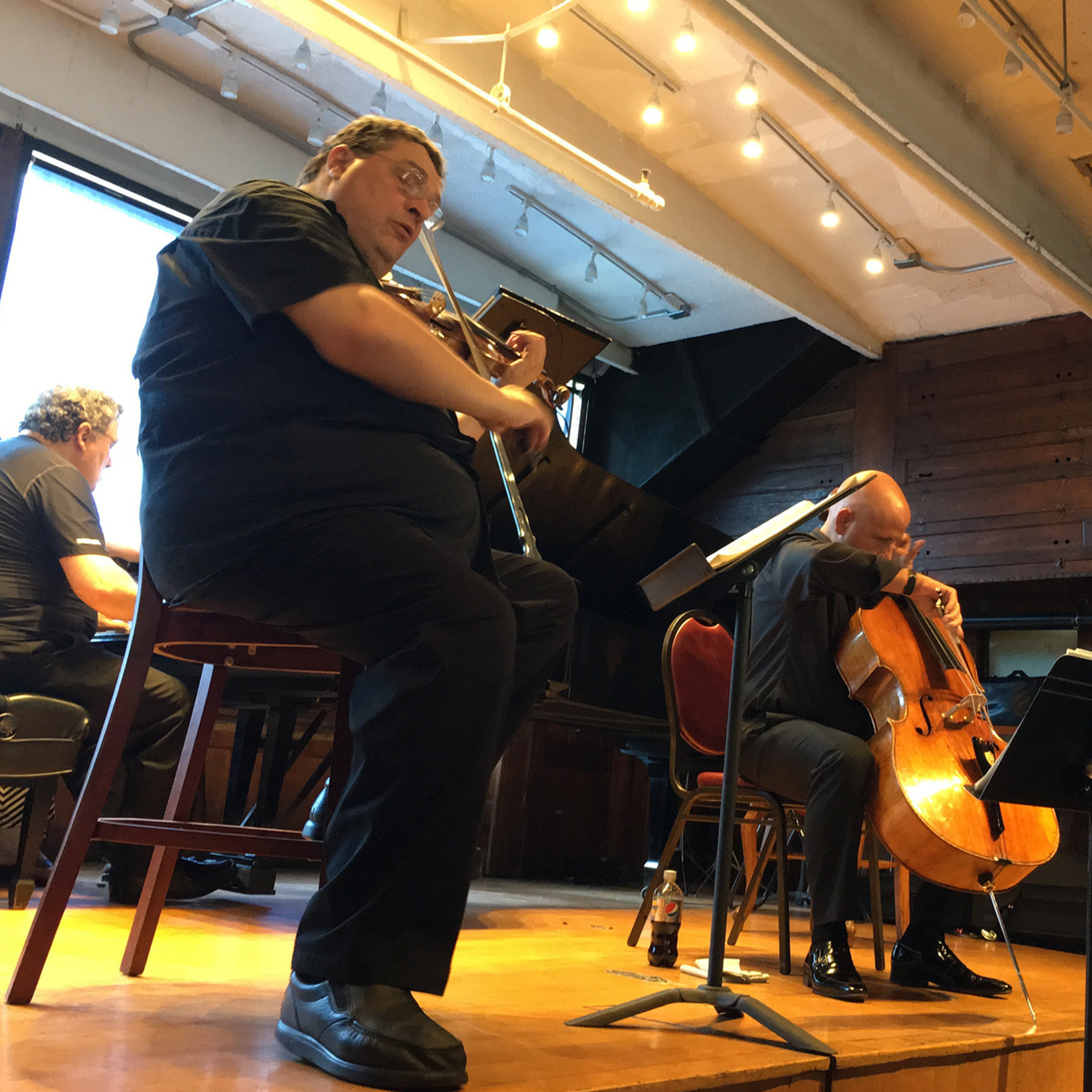 Three musicians performing chamber music in a cozy, wood-paneled venue, violin, cello, and piano