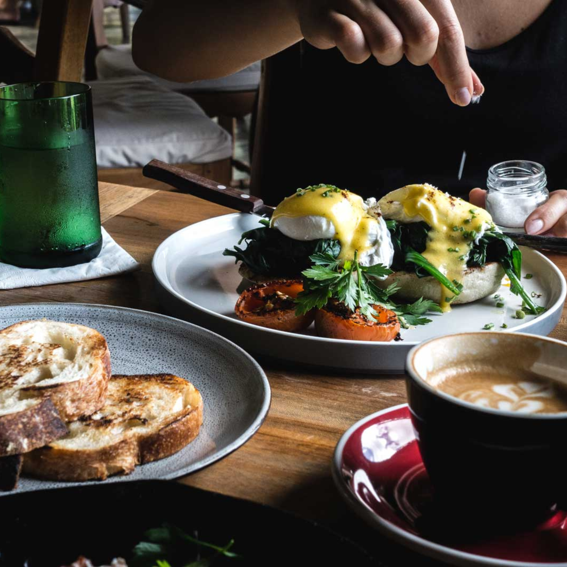 Person sprinkling salt on Eggs Benedict at a cozy café table with toast, coffee, and greens