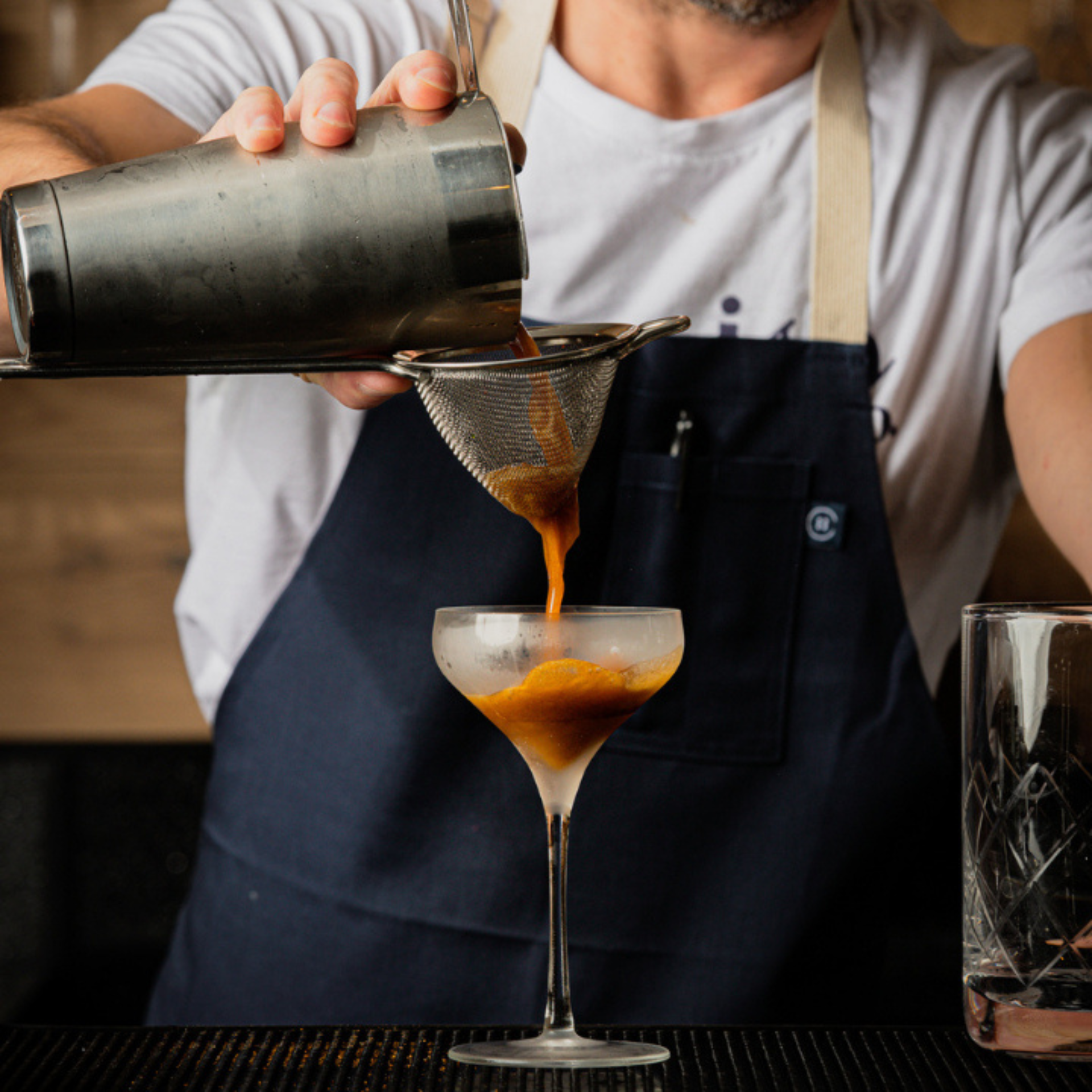 Bartender straining dark cocktail into coupe glass at bar
