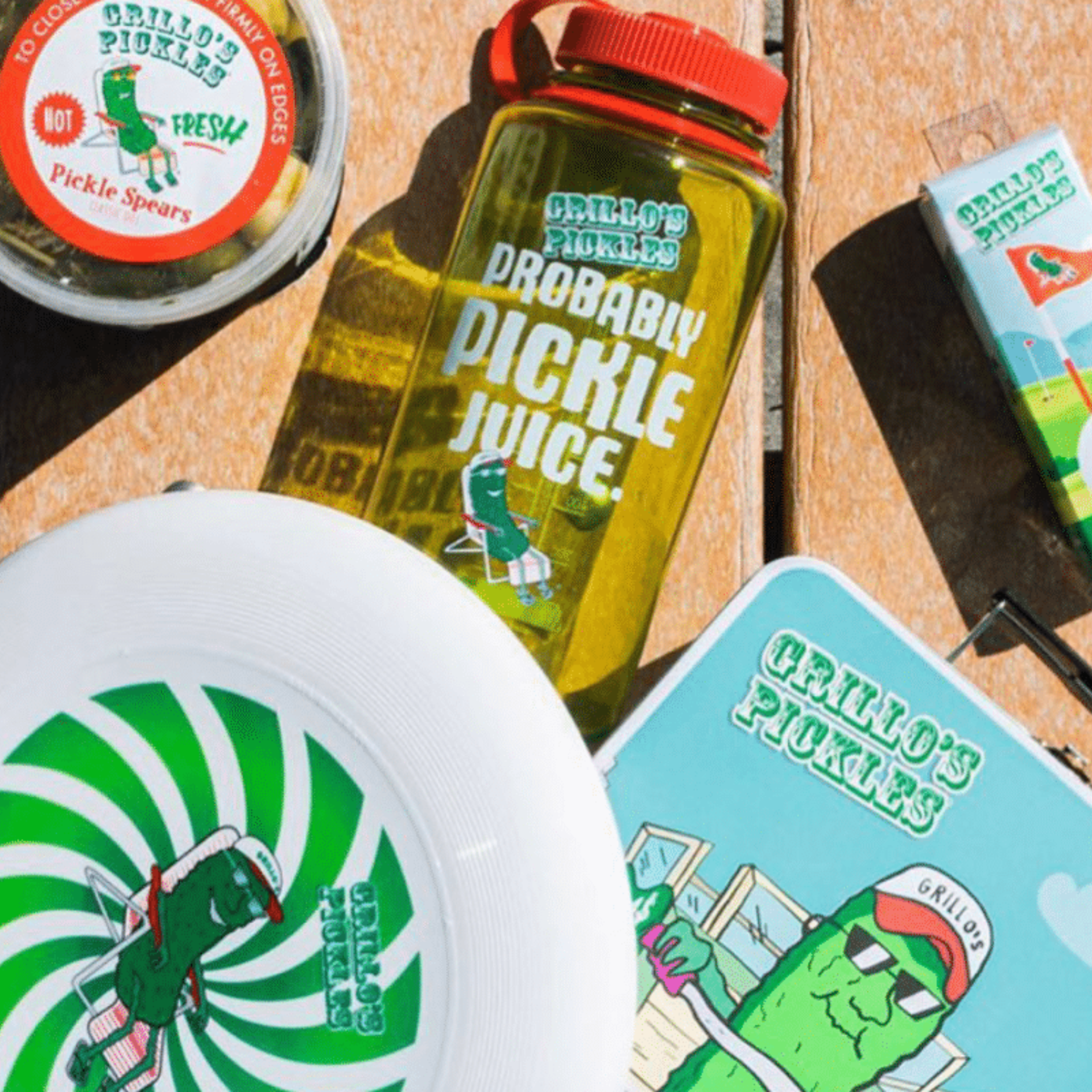Assorted Grillo’s Pickles merchandise including a pickle container, water bottle, frisbee, and tin with cartoon pickle designs
