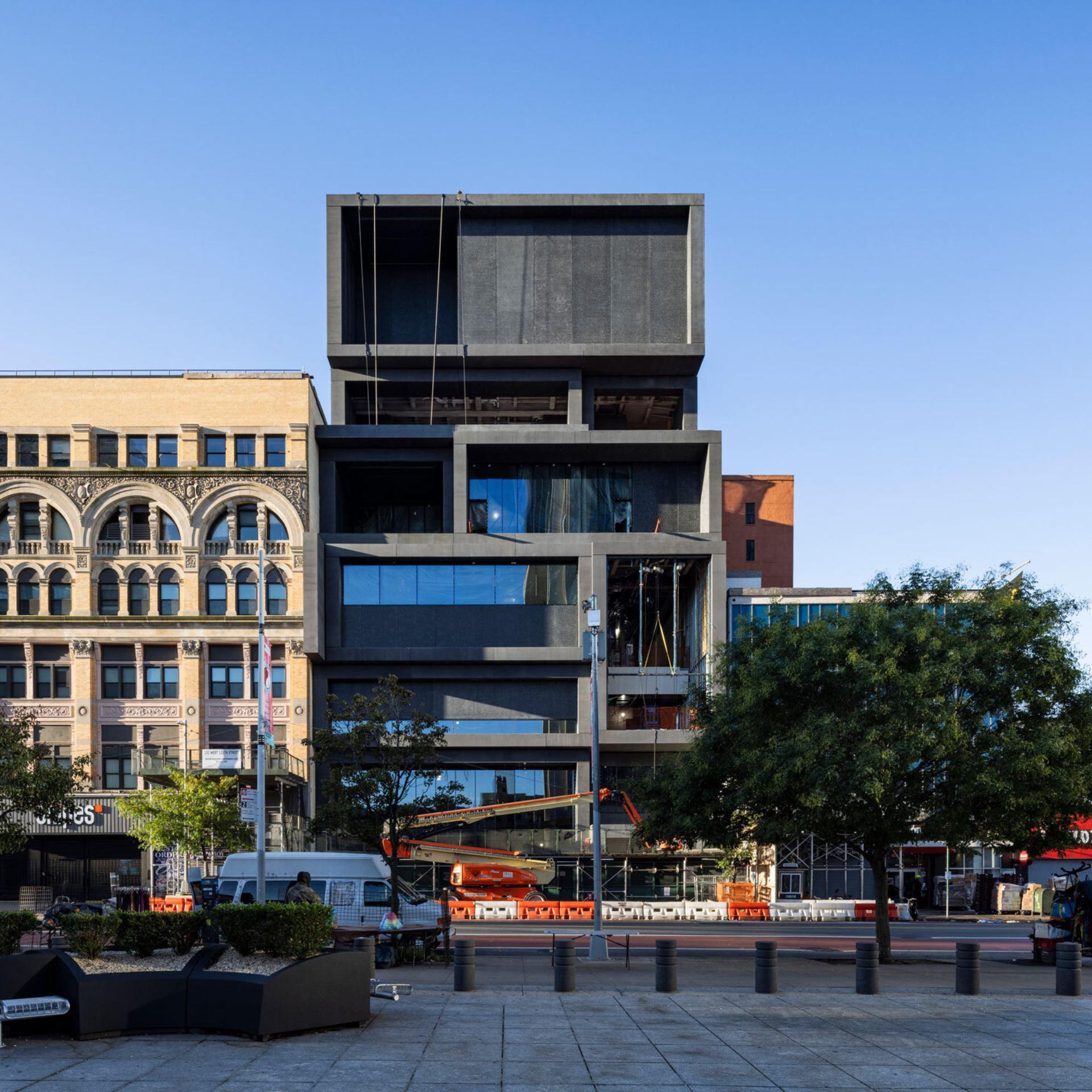 Modern dark-toned building with offset stacked levels, under construction beside older ornate structures in a city plaza