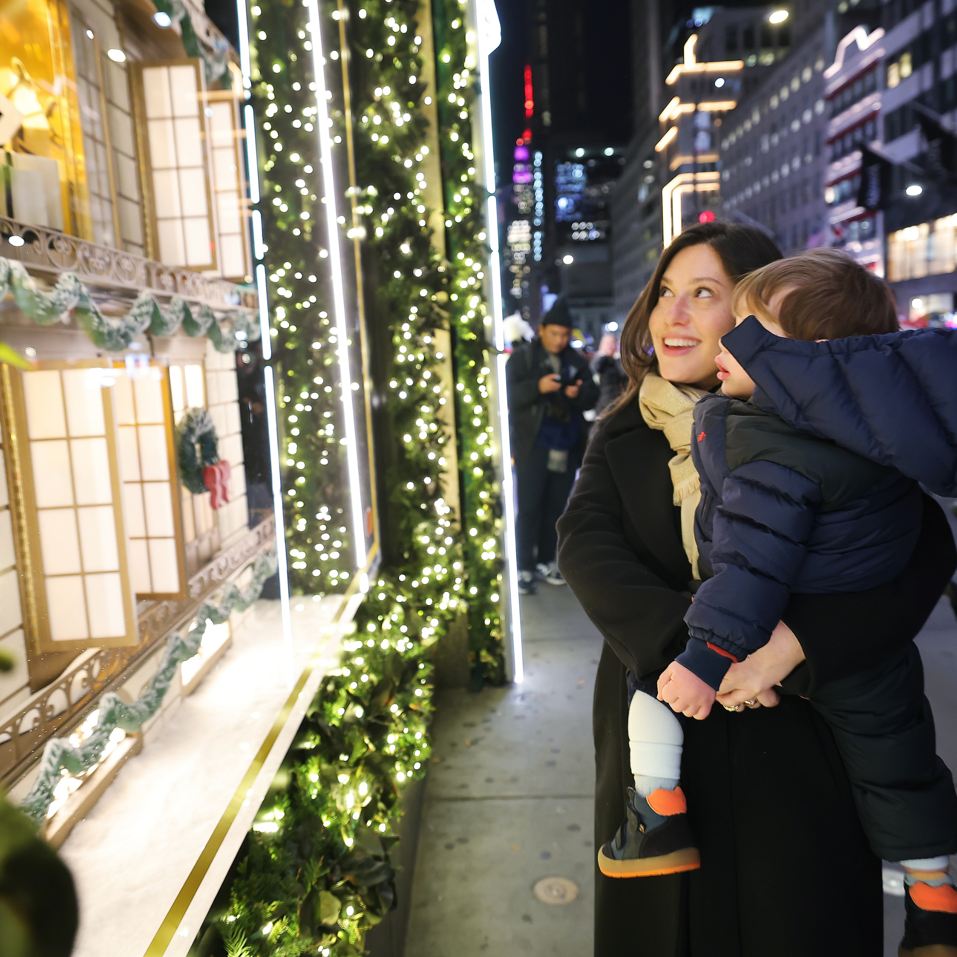 Woman and child admiring a festive city window display with miniature buildings and holiday lights at night