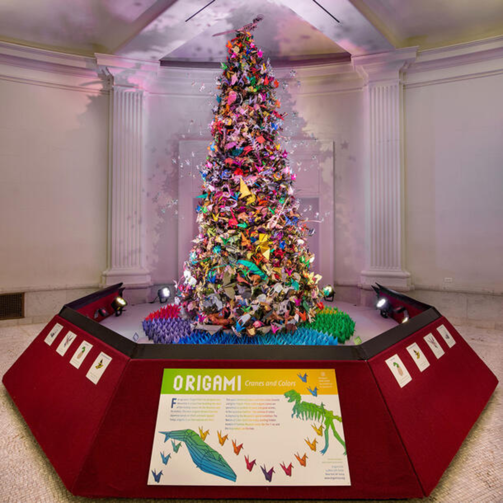 Christmas tree decorated with colorful origami figures, displayed under spotlights in a classical room