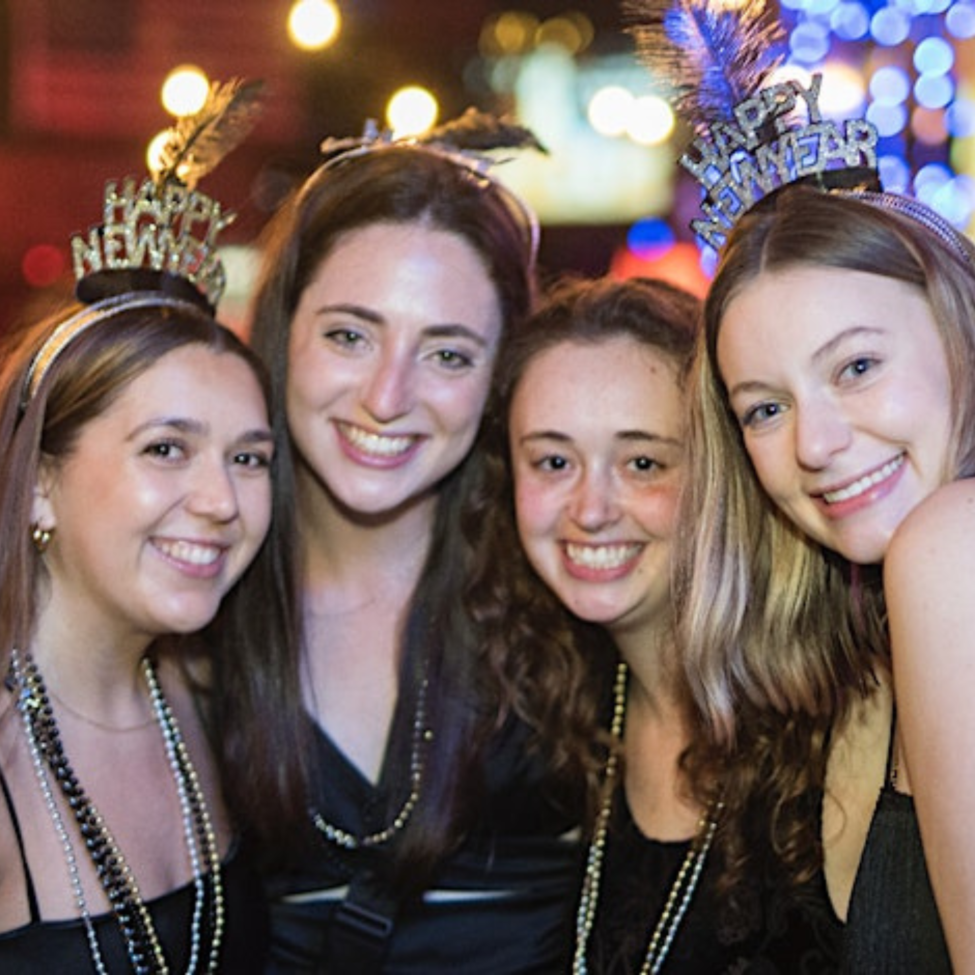Four people wearing 'Happy New Year' headbands and beaded necklaces, smiling together at a nighttime celebration