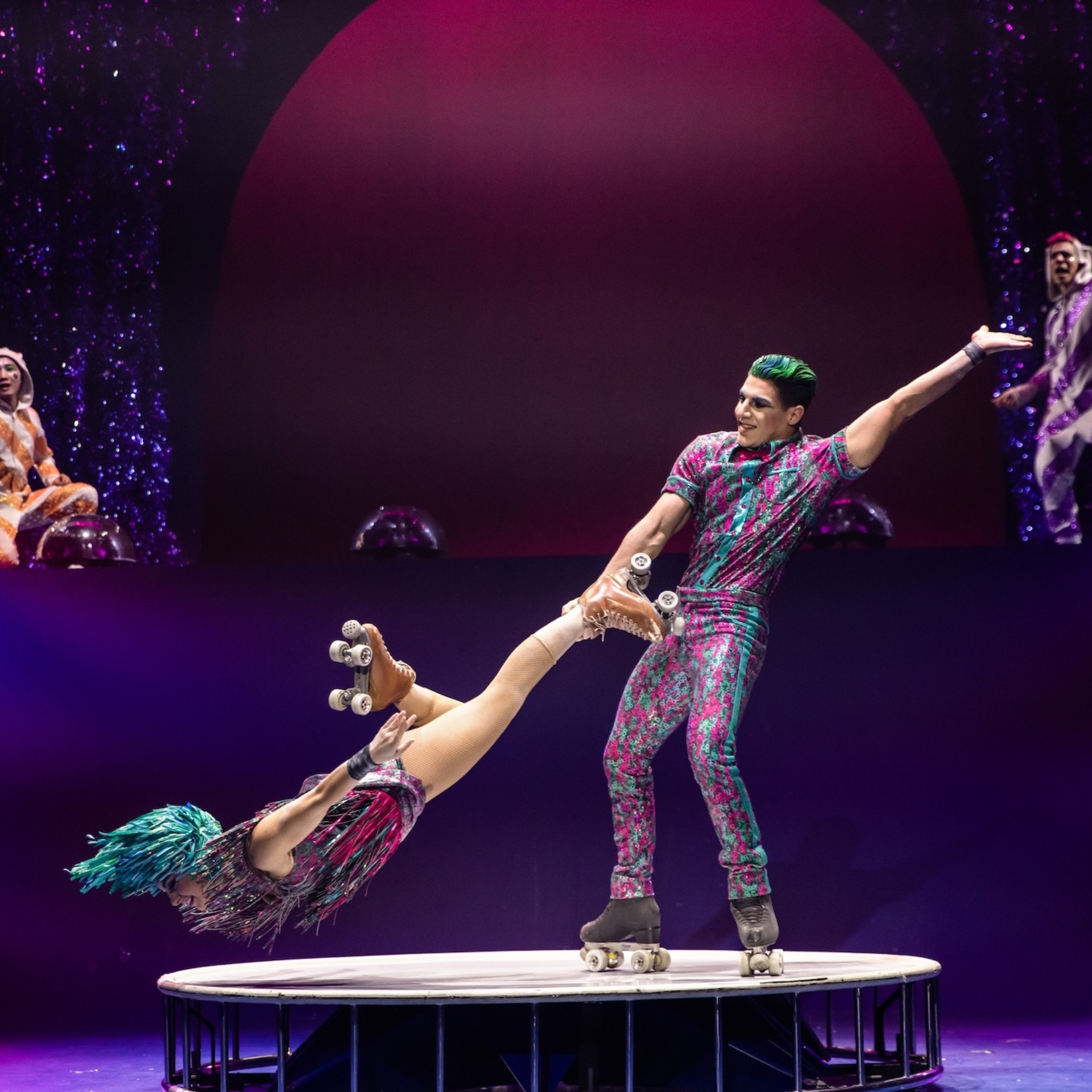 Roller skaters performing an acrobatic stunt on a circular stage, surrounded by colorful costumes and glittering set