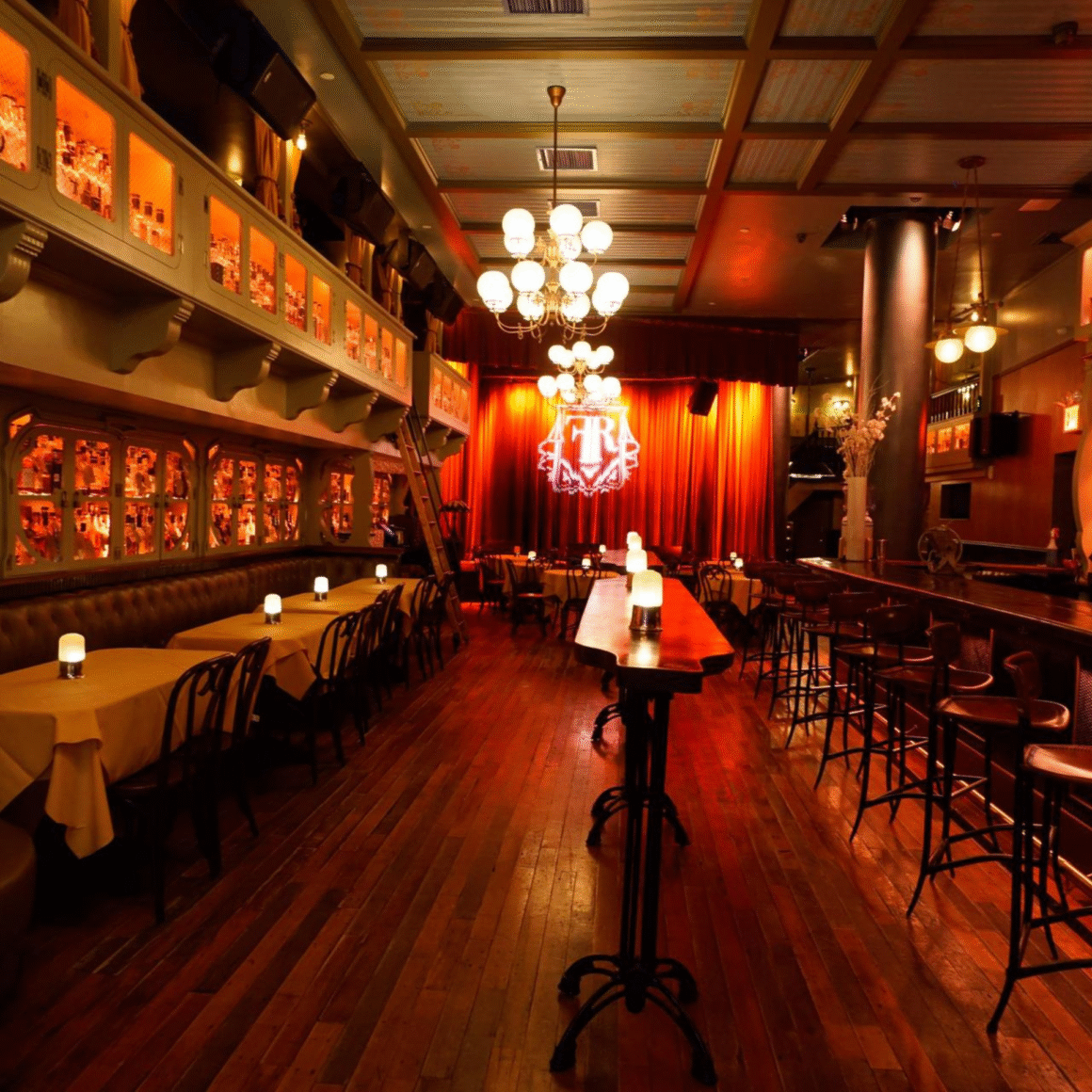 Elegant lounge-style restaurant with warm lighting, wooden floors, set tables, and a red curtain stage at the back