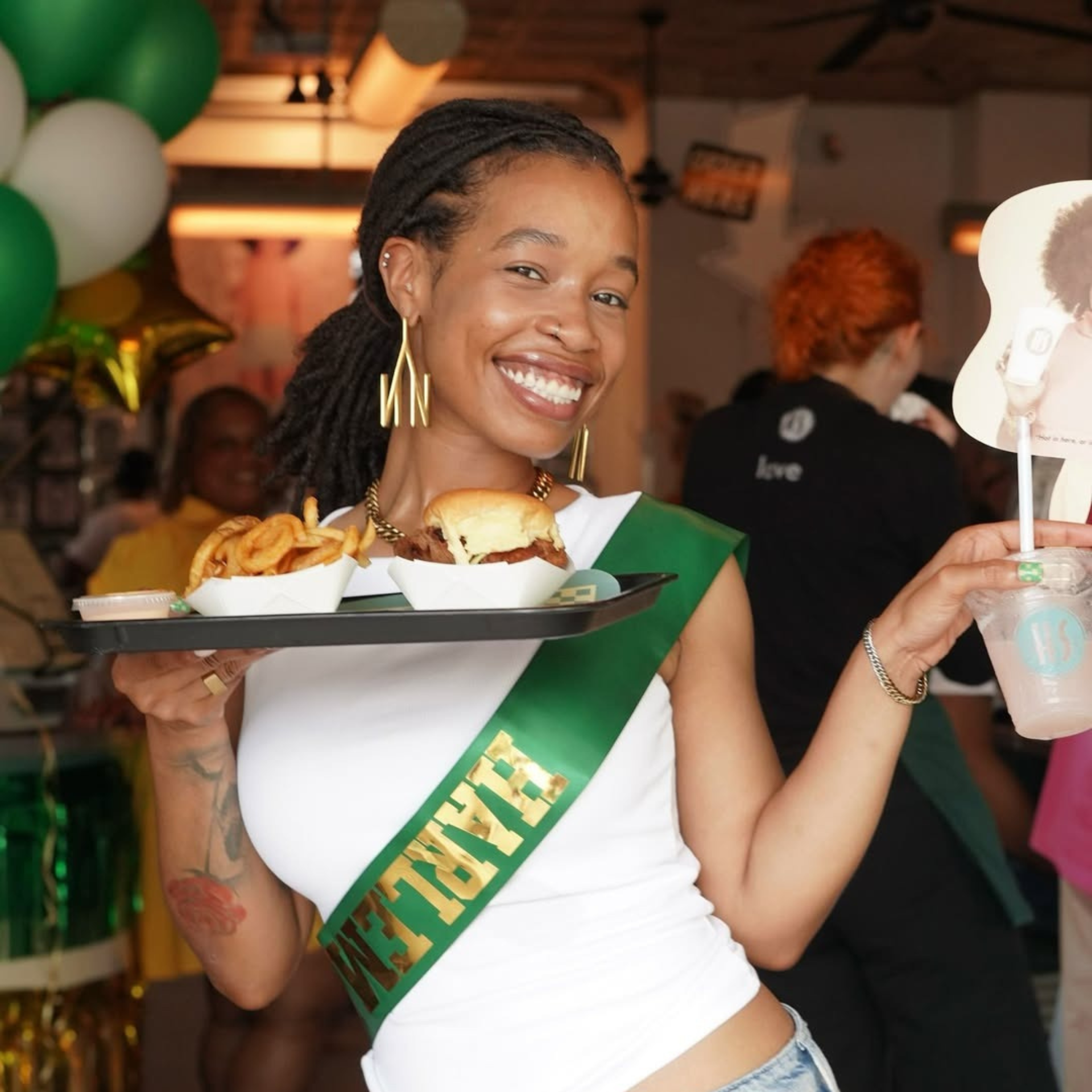 Smiling person holding a tray of food and drink at a festive indoor event with balloons and decorations