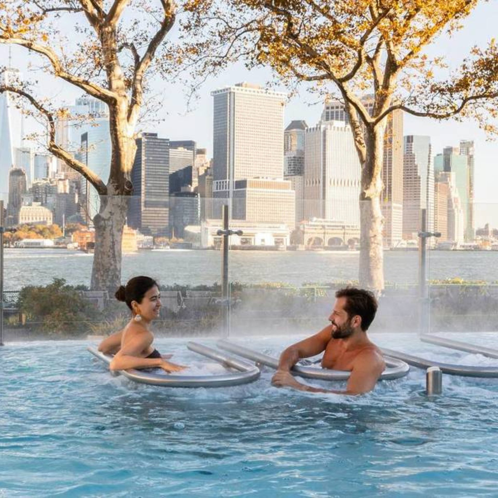 Two people relaxing in a heated outdoor pool with city skyline views and autumn trees in the background