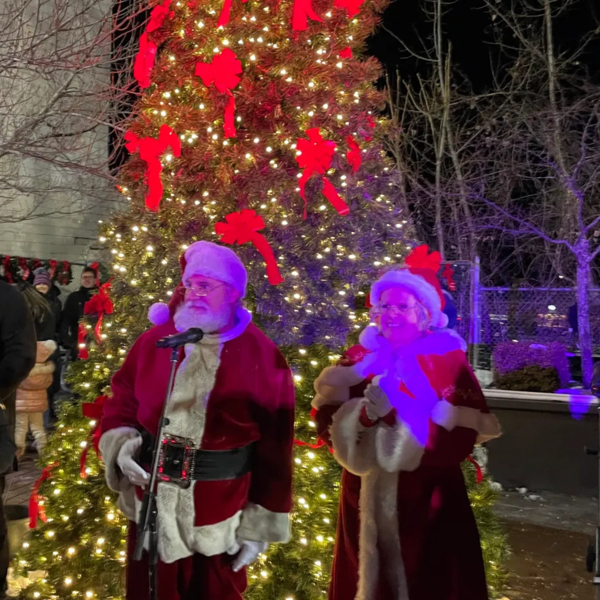 Santa and Mrs. Claus standing in front of a lit Christmas tree at a festive outdoor nighttime event