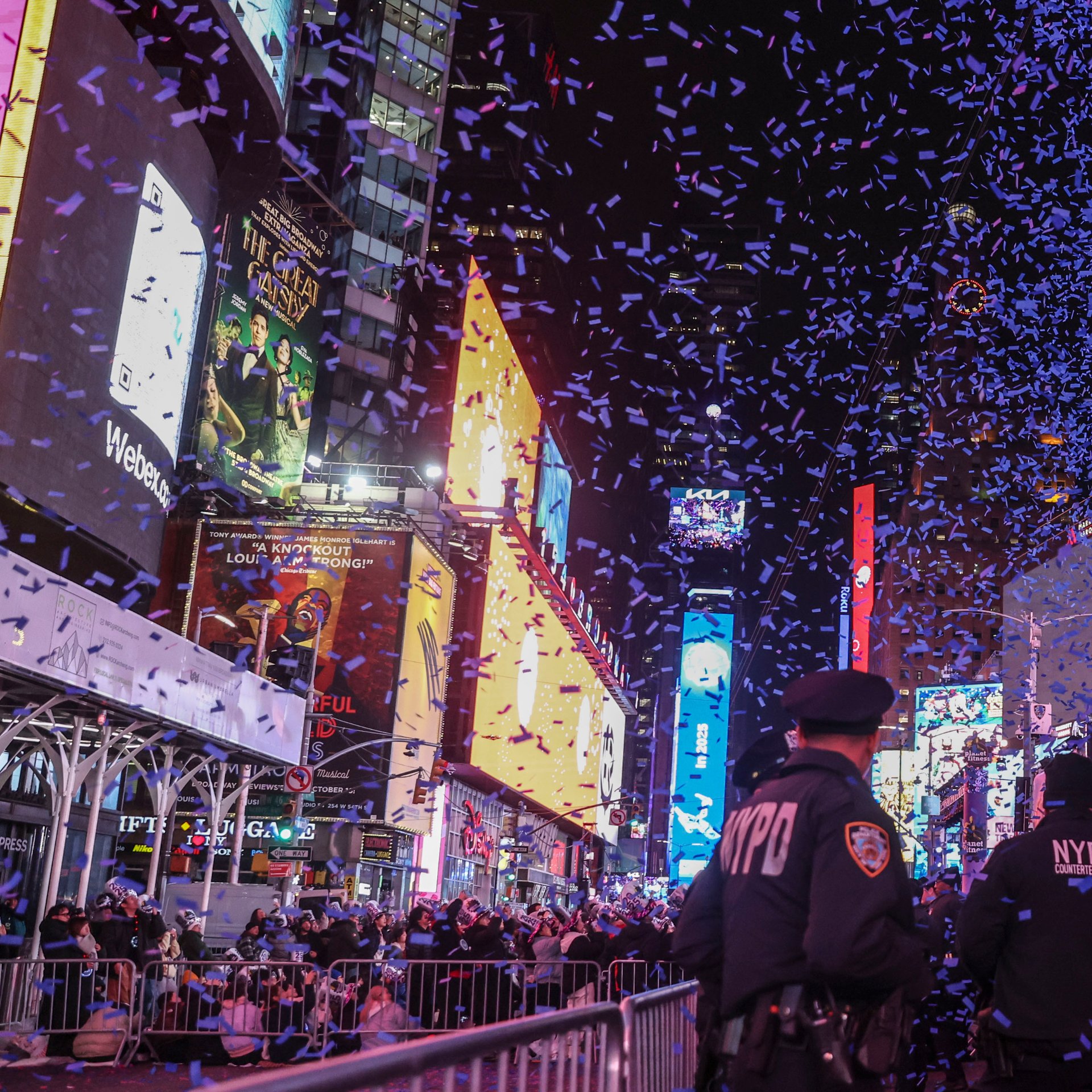 Crowds celebrating in Times Square at night with falling confetti, bright billboards, and NYPD officers