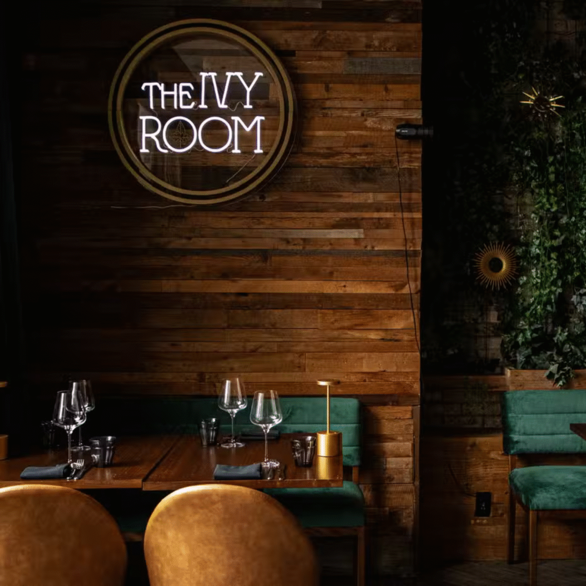 Cozy restaurant interior with a wooden wall, green seating, set dining table, and a glowing “The Ivy Room” sign