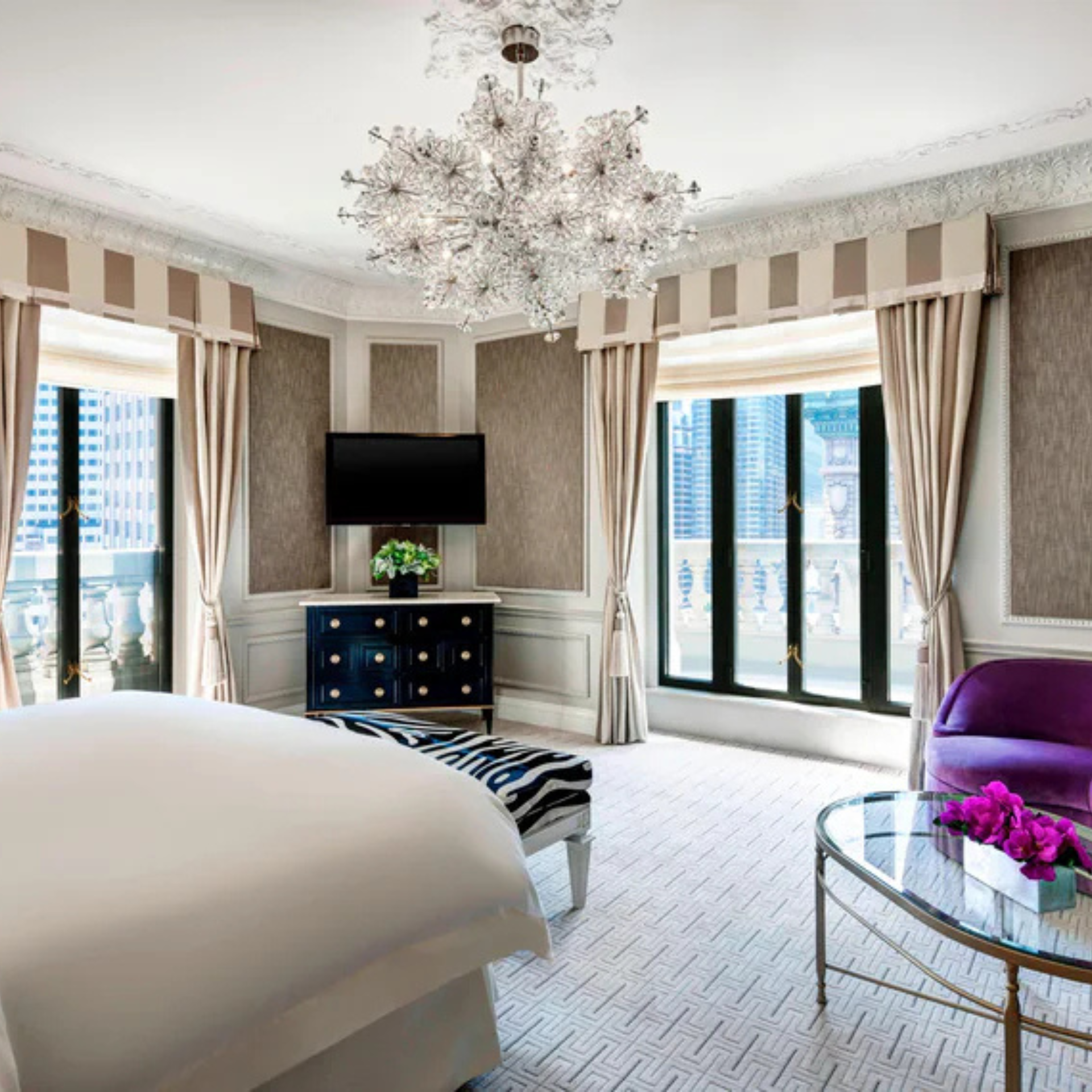 Luxurious hotel room with white linens, zebra-patterned bench, purple accents, and city view windows