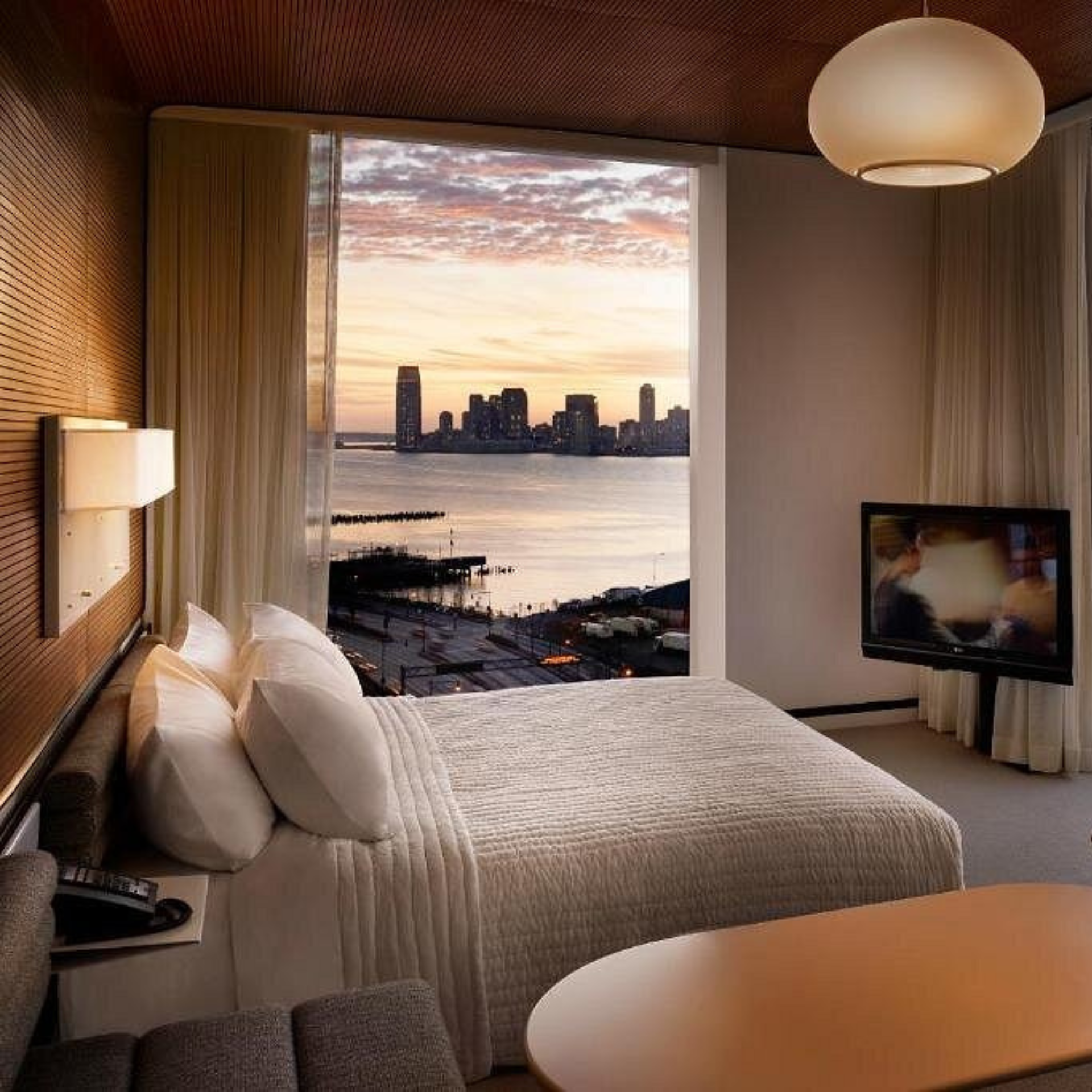 Modern hotel room with white linens, sunset city view through large window, and sleek furnishings