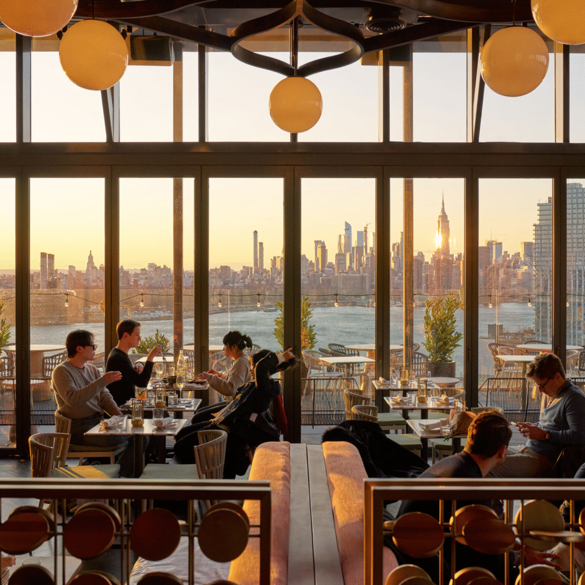 People dining inside a modern restaurant with floor-to-ceiling windows overlooking the New York City skyline at sunset